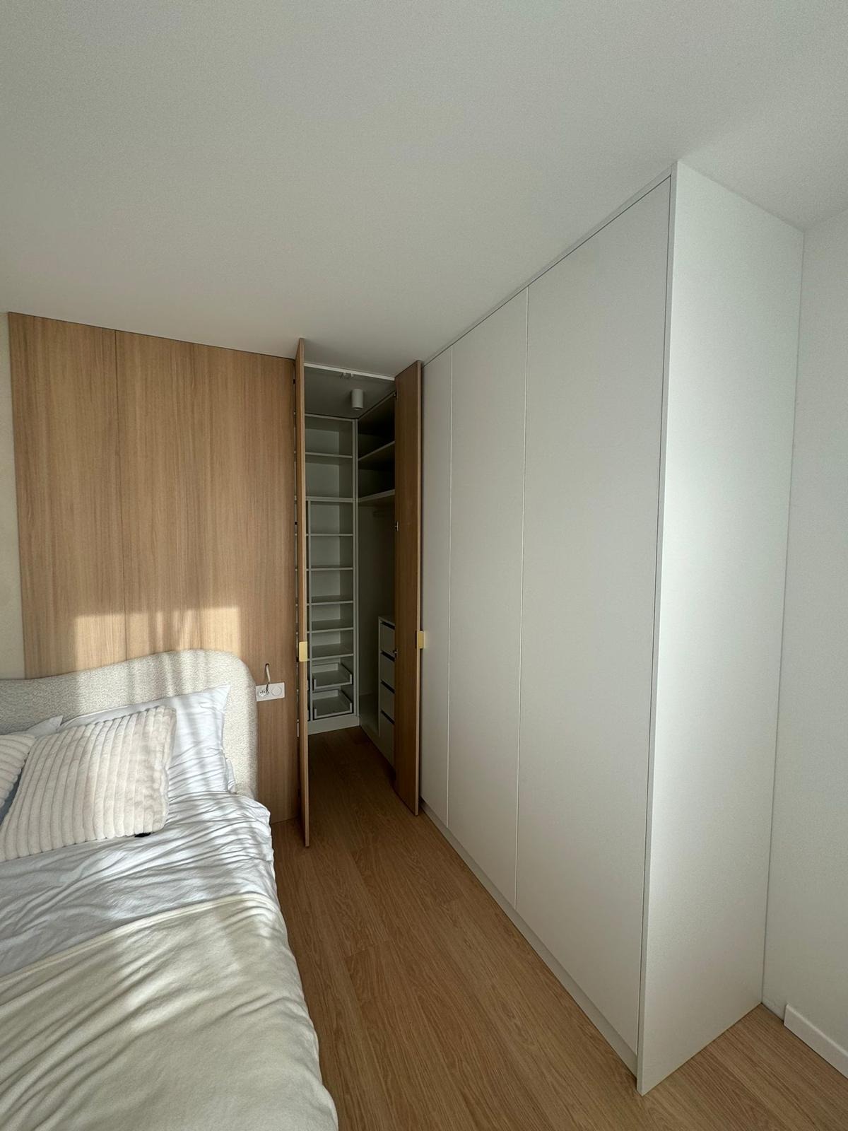 Nowoczesna szafa wnękowa z białymi frontami, częściowo otwarta, z drewnianym wykończeniem obok łóżka w sypialni.