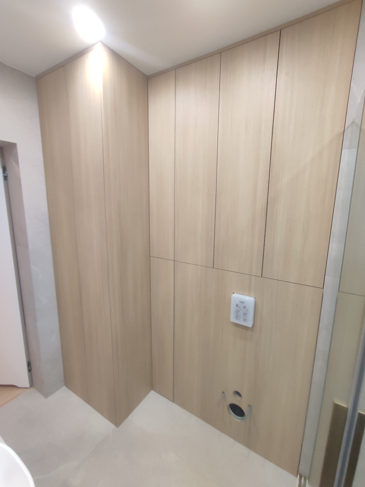 Jasne, drewniane szafy wnękowe w łazience z widocznym panelem sterowania i otworem wentylacyjnym w dolnej części.