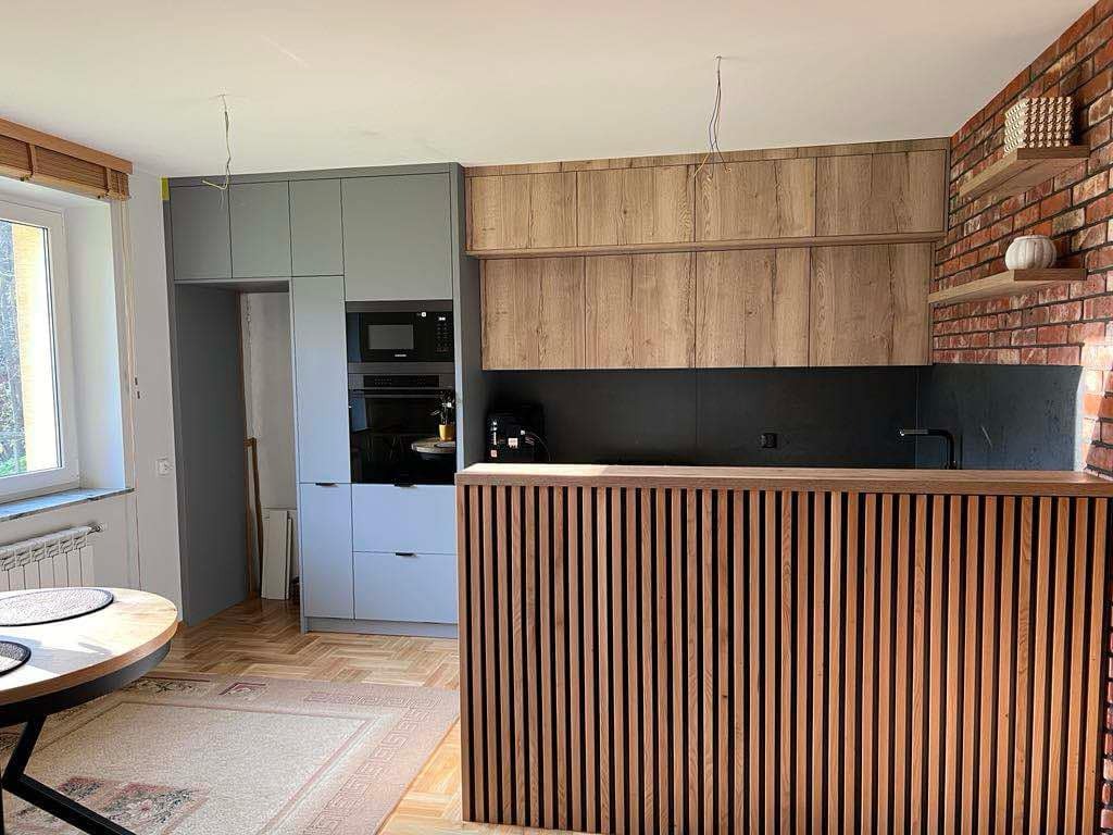 Nowoczesna kuchnia z szarymi i drewnianymi szafkami, czarnym blatem i ceglaną ścianą z półkami, widok na okrągły stół i dywan.