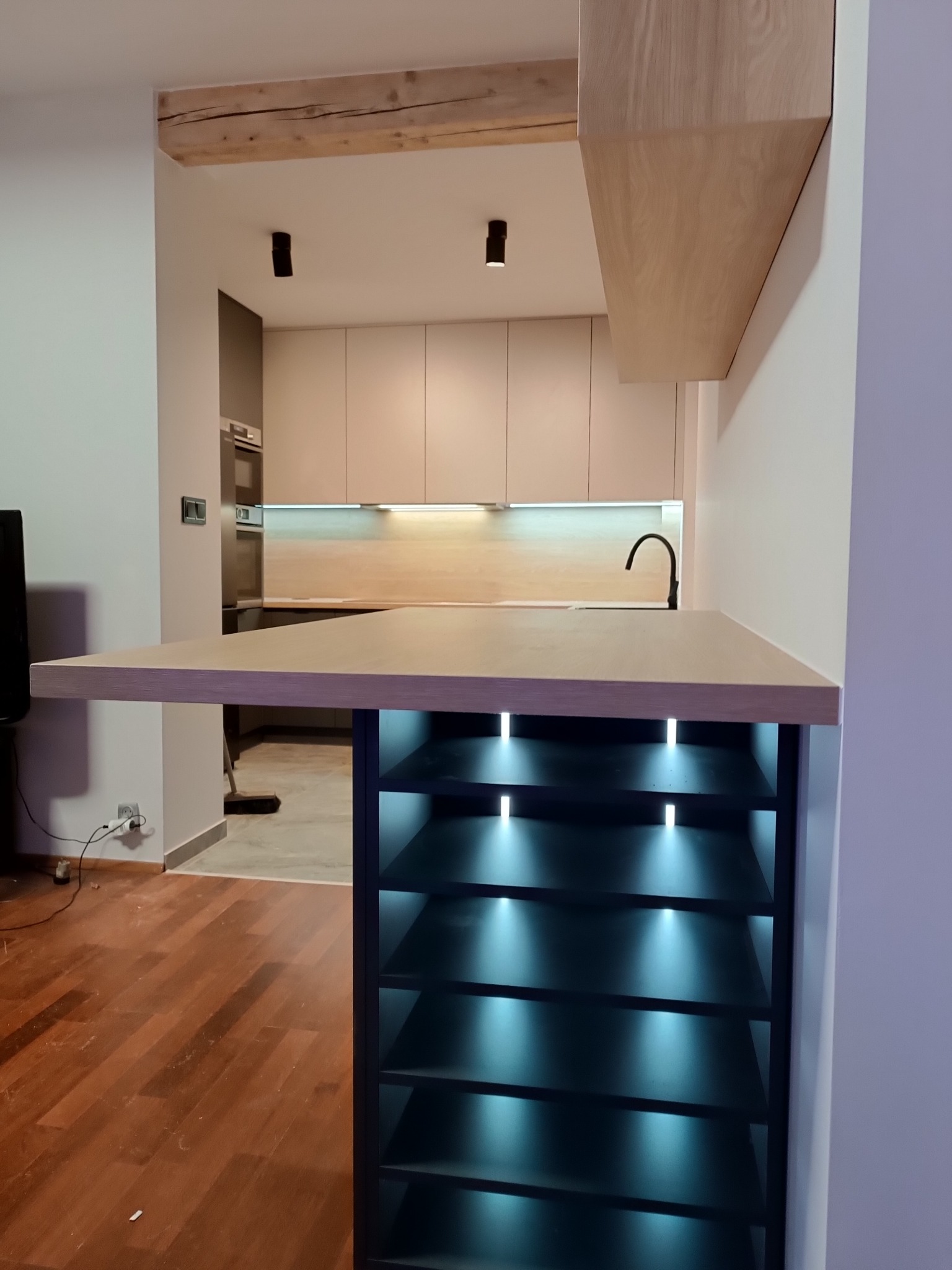 Nowoczesna kuchnia z blatem barowym z podświetlanymi półkami, widoczne drewniane elementy wykończenia i czarna bateria.