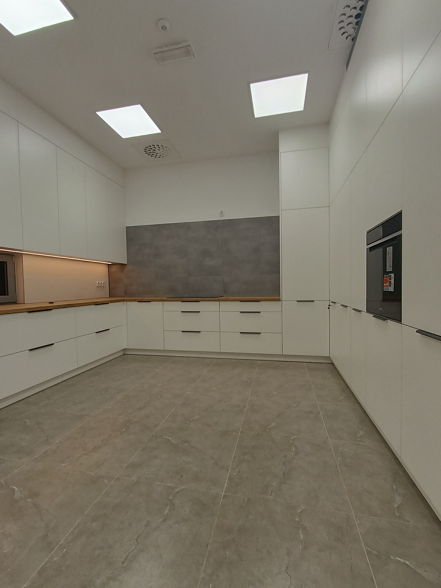 Minimalistyczna, nowoczesna kuchnia z białymi frontami szafek i drewnianym blatem, szara ściana nad blatem, oświetlenie LED pod szafkami, piekarnik w zabudowie, płytki podłogowe imitujące kamień.