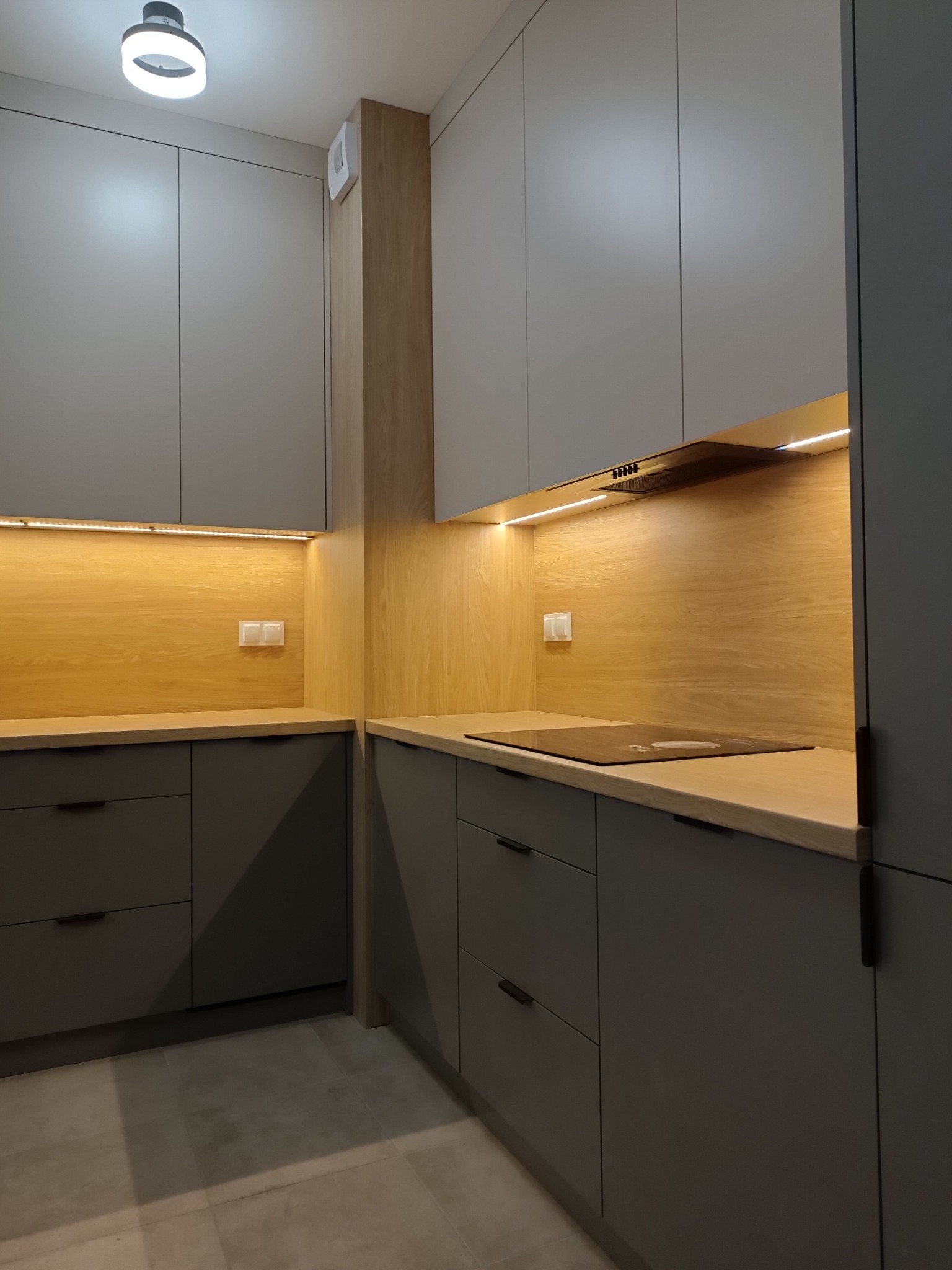 Szare matowe szafki kuchenne z drewnianym panelem ściennym i oświetleniem LED podszafkowym, widoczna płyta indukcyjna i minimalistyczne uchwyty.