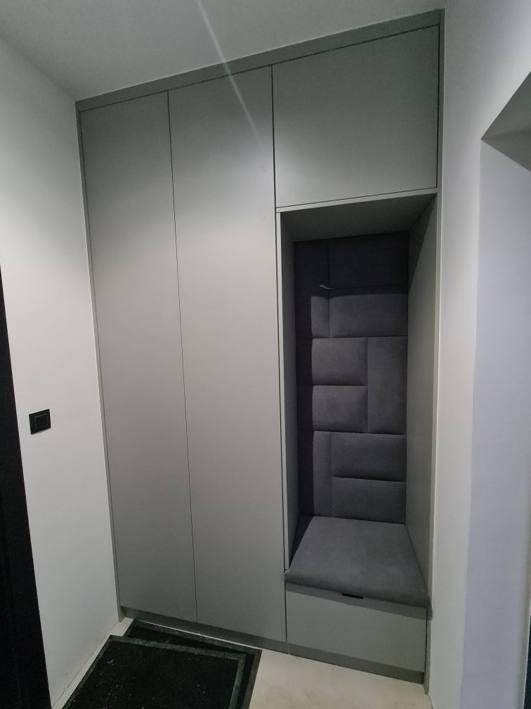 Szara szafa wnękowa z siedziskiem obitym ciemnoszarą tapicerką z geometrycznym wzorem na ścianie wnęki.