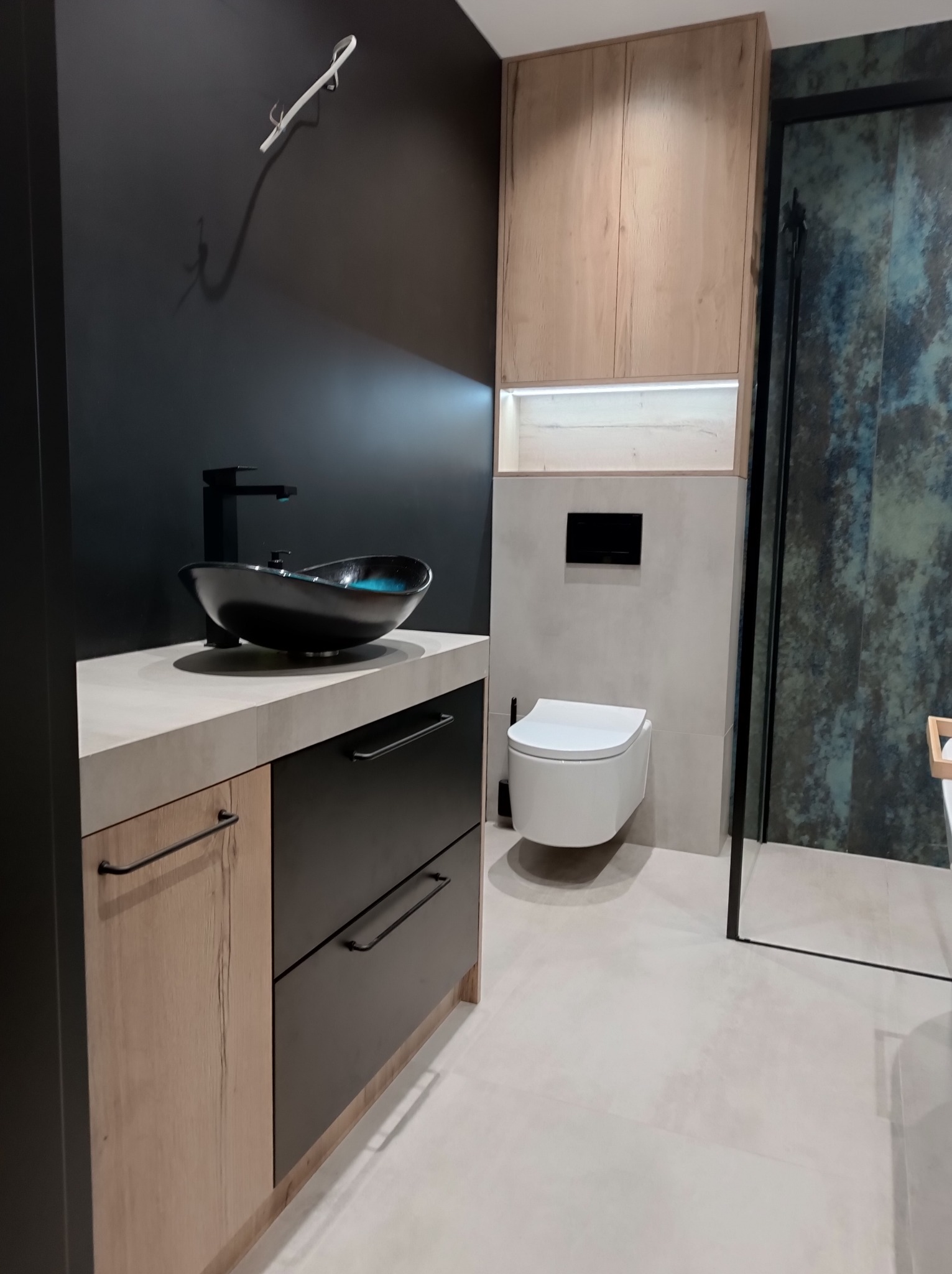 Nowoczesna łazienka z umywalką nablatową w kształcie łodzi, czarną armaturą, szafką z frontami drewnopodobnymi i czarnymi szufladami, toaletą podwieszaną i kabiną prysznicową z dekoracyjną ścianą.