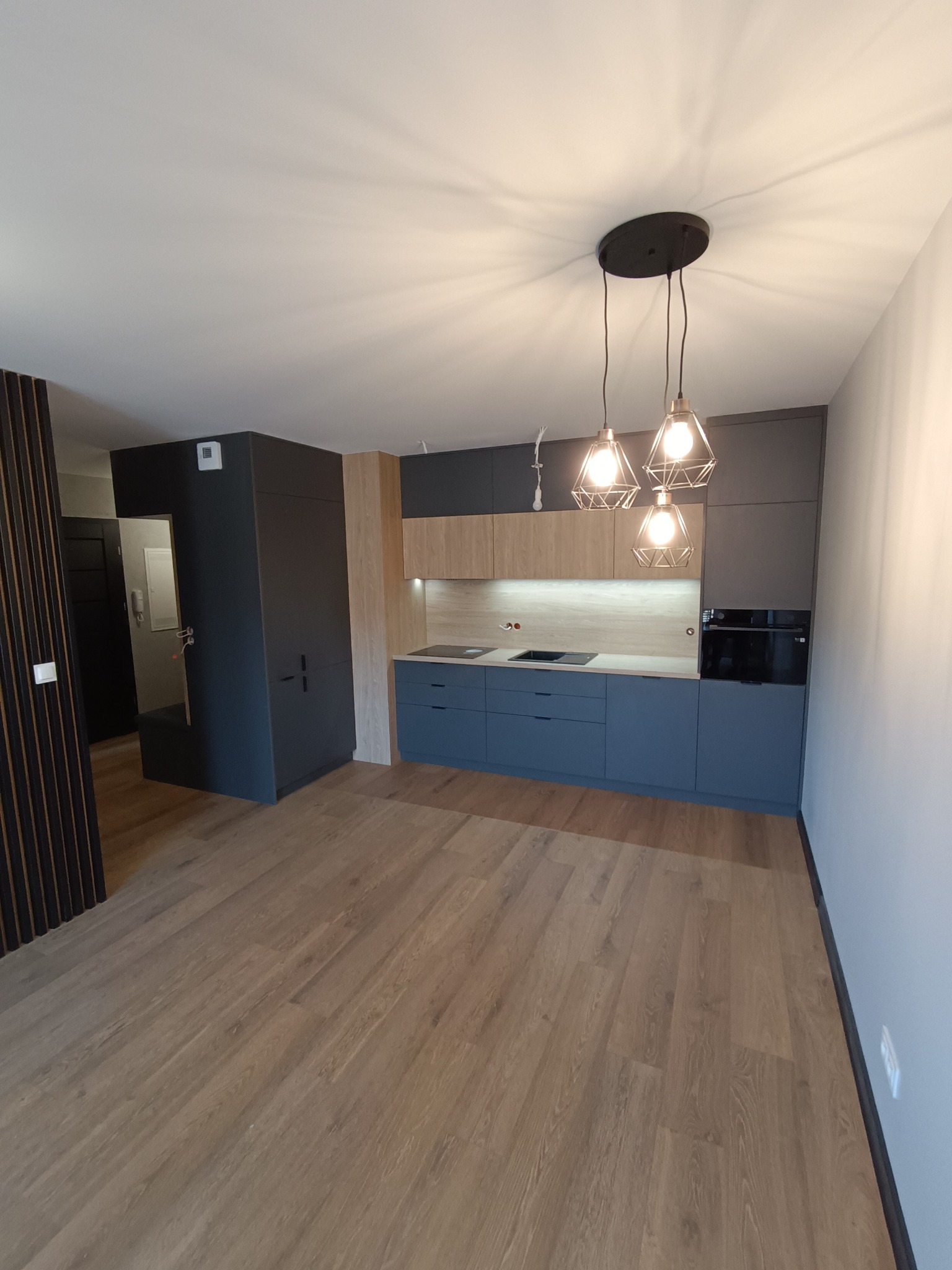 Nowoczesna kuchnia z niebieskimi szafkami dolnymi, drewnianymi górnymi i trzema lampami wiszącymi o geometrycznych kloszach nad blatem.