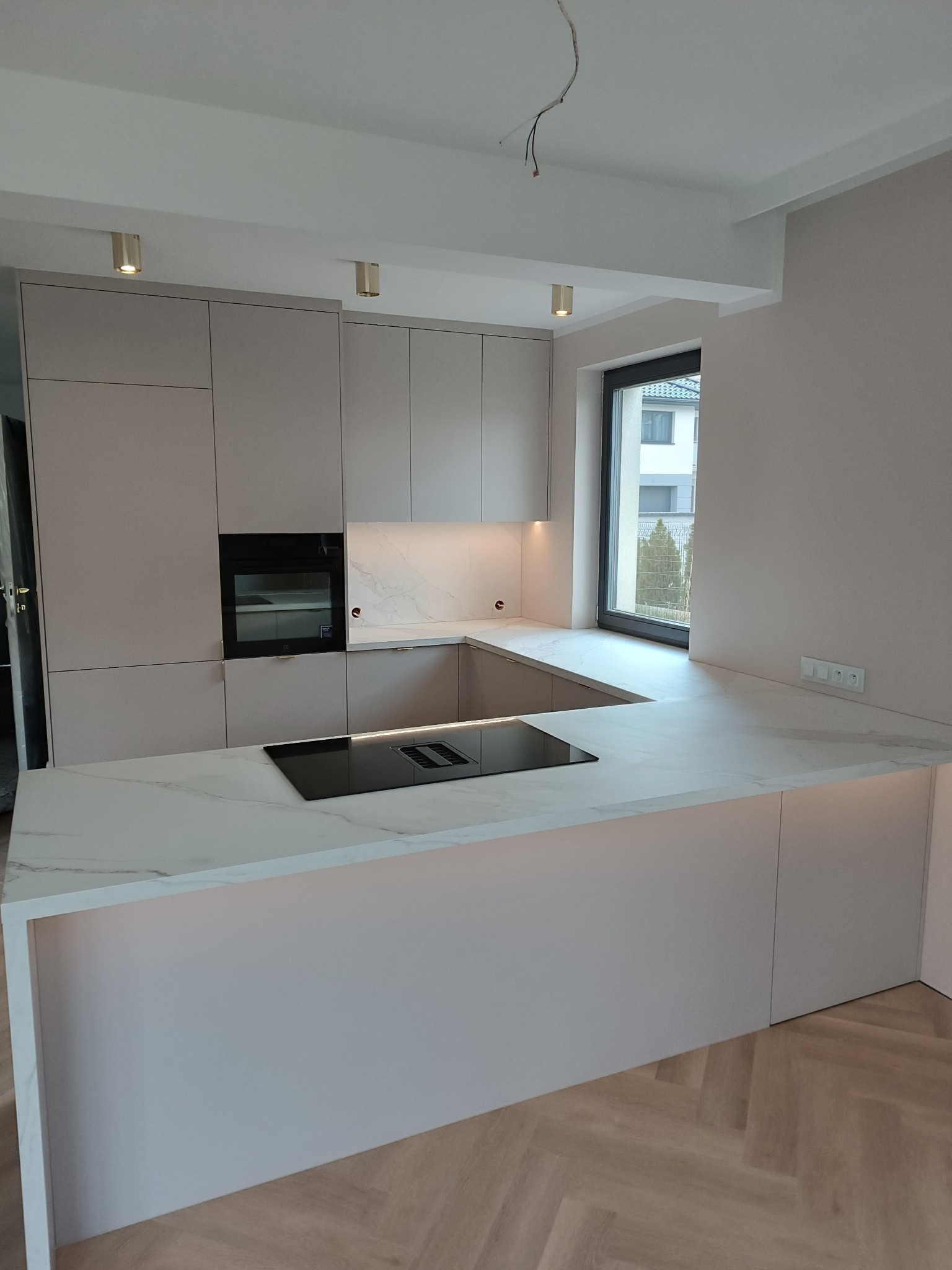 Jasna kuchnia na wymiar z marmurowym blatem i wyspą kuchenną z wbudowaną płytą indukcyjną, oświetleniem podszafkowym i parkietem ułożonym w jodełkę.