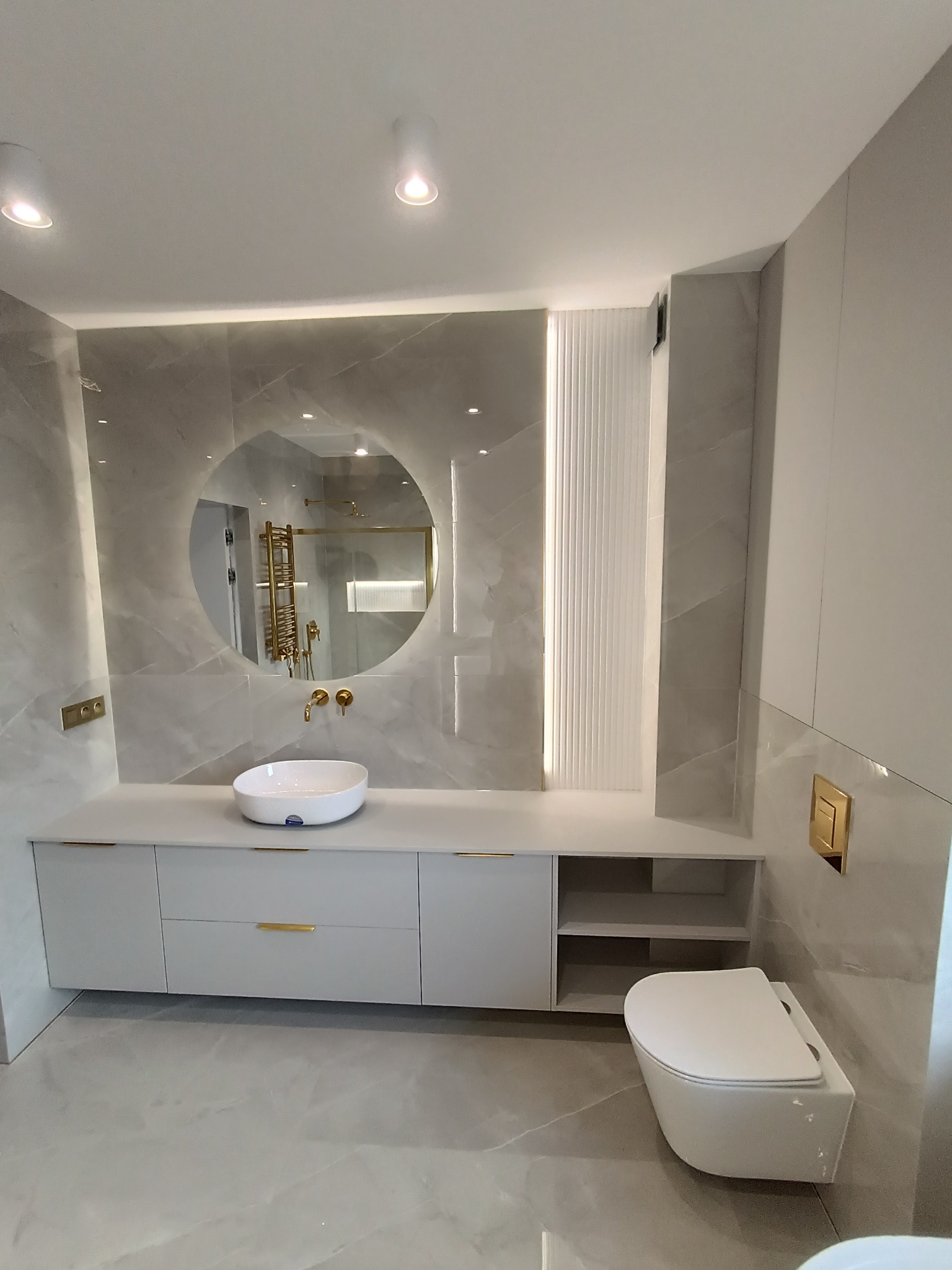 Nowoczesna łazienka z szarą zabudową szafek pod umywalką, okrągłym lustrem z podświetleniem LED, złotymi akcentami w armaturze i elementach wykończeniowych, oraz podwieszaną toaletą.