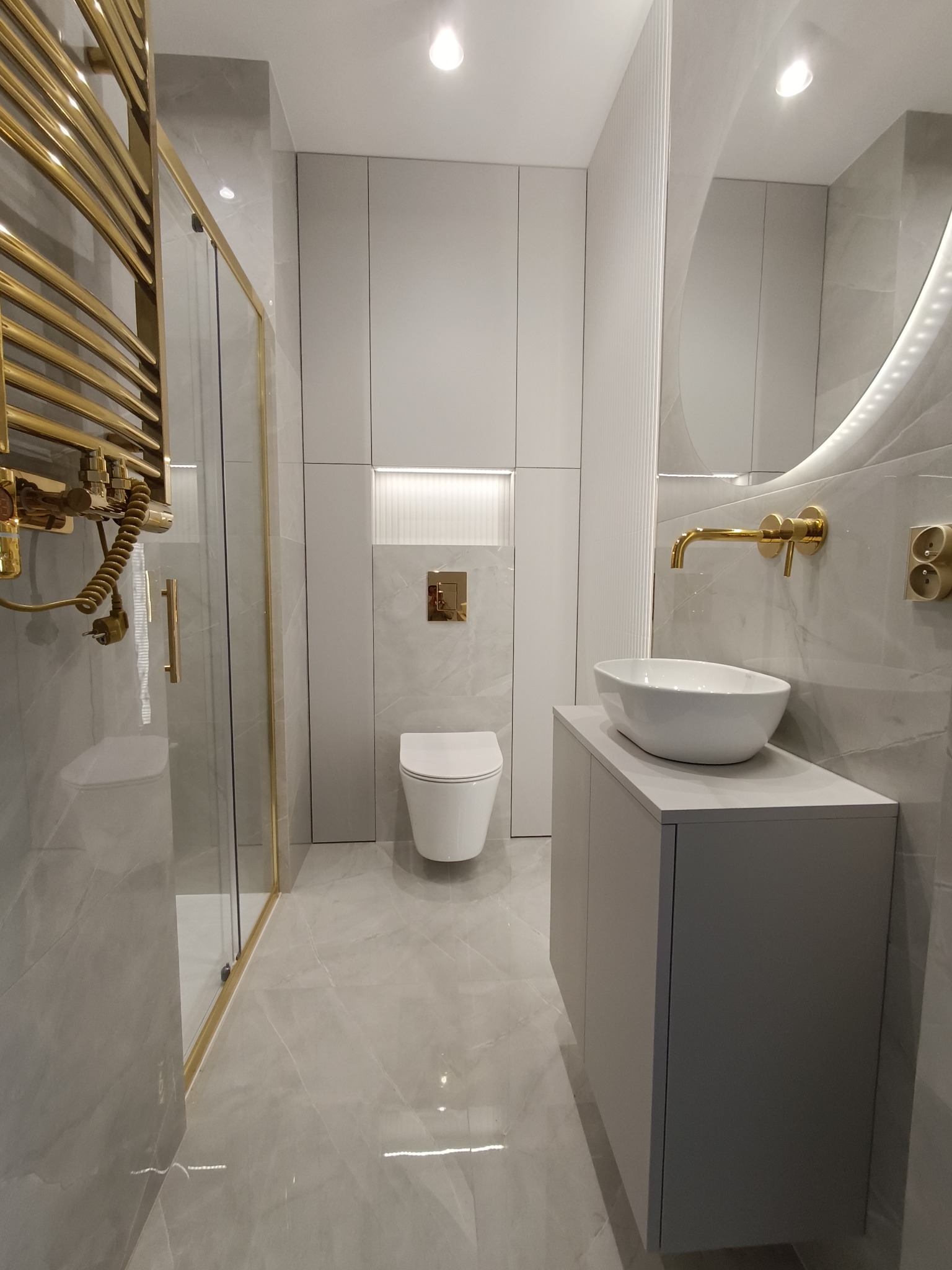 Nowoczesna łazienka w odcieniach szarości i złota: kabina prysznicowa ze złotymi ramami, grzejnik drabinkowy w kolorze złota, wisząca toaleta, umywalka nablatowa na szarej szafce oraz okrągłe...