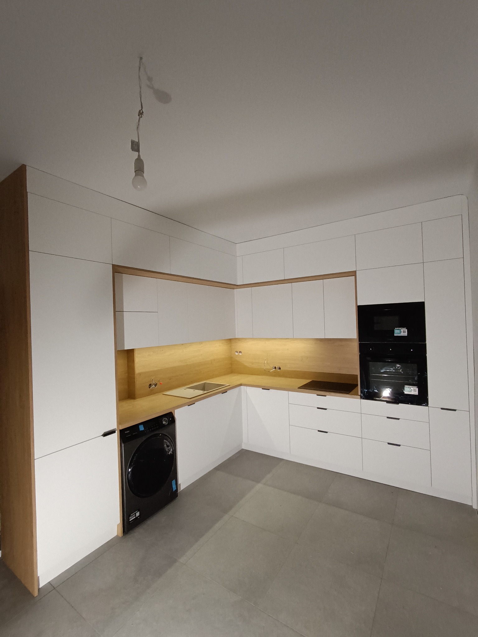 Nowoczesna kuchnia z białymi frontami i drewnianymi akcentami, oświetleniem LED pod szafkami wiszącymi, z wbudowaną pralką i piekarnikiem w zabudowie, surowa żarówka zwisająca z sufitu.