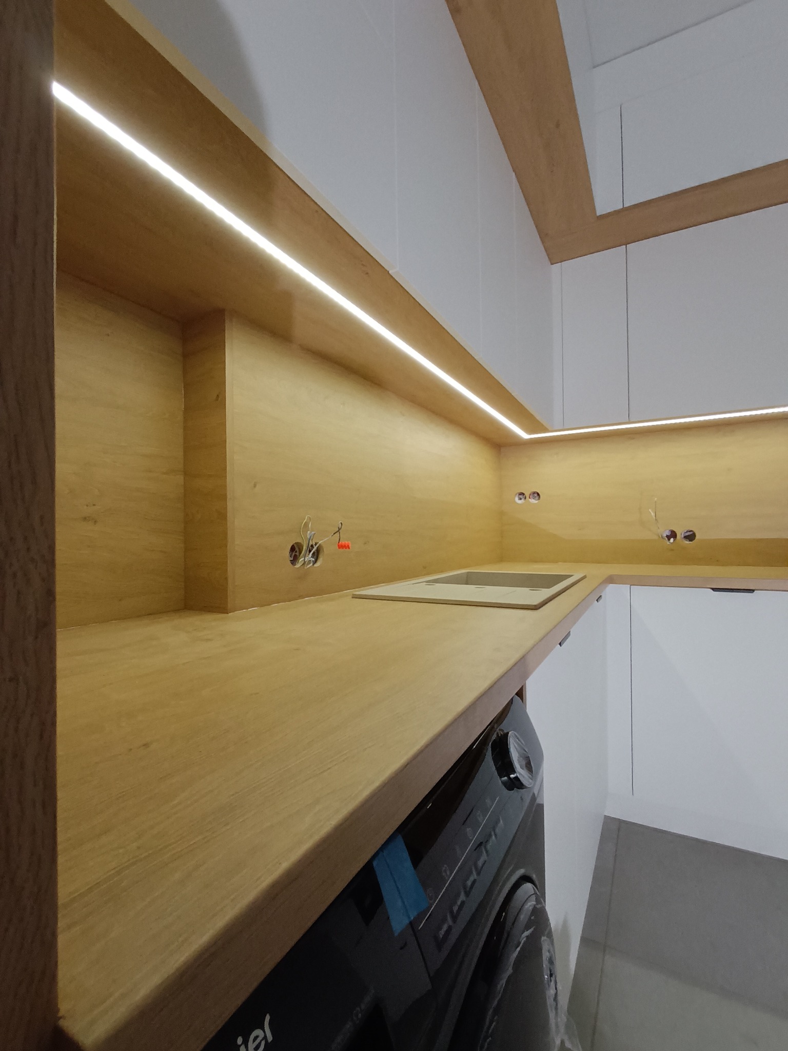 Nowoczesna kuchnia z drewnianymi blatami, białymi szafkami i oświetleniem LED podszafkowym; widoczna pralka pod blatem, instalacja elektryczna w ścianie.