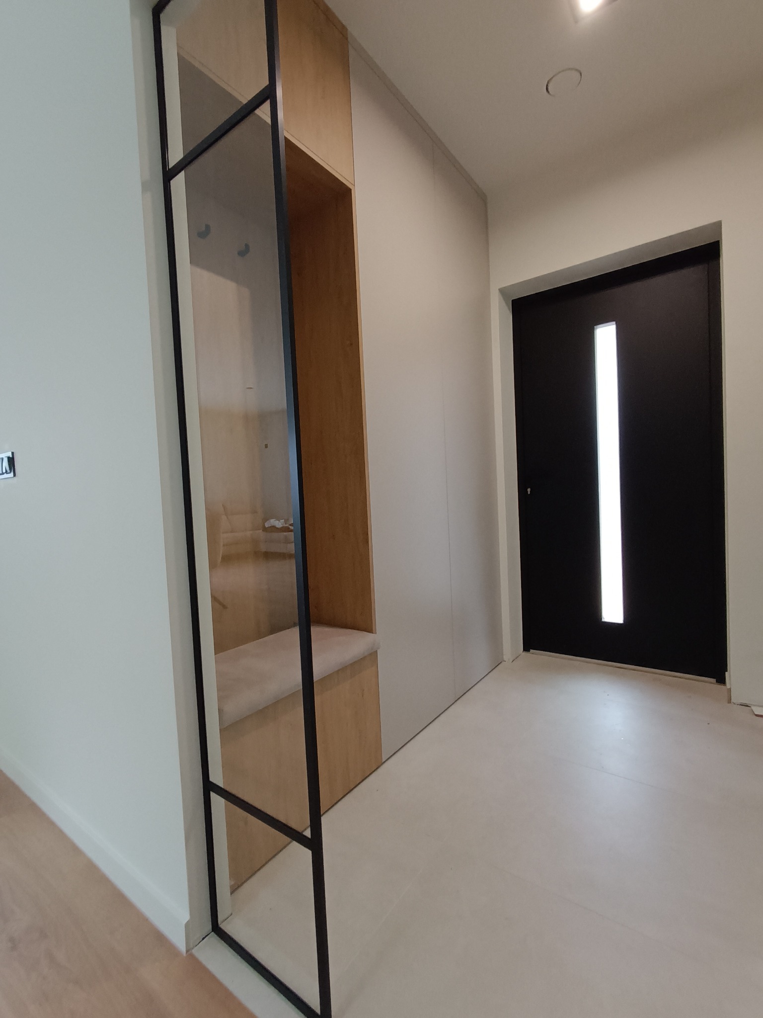 Nowoczesny przedpokój z zabudowaną szafą wnękową z siedziskiem i czarnymi, metalowymi elementami wykończeniowymi, widok na czarne drzwi wejściowe z przeszkleniem.