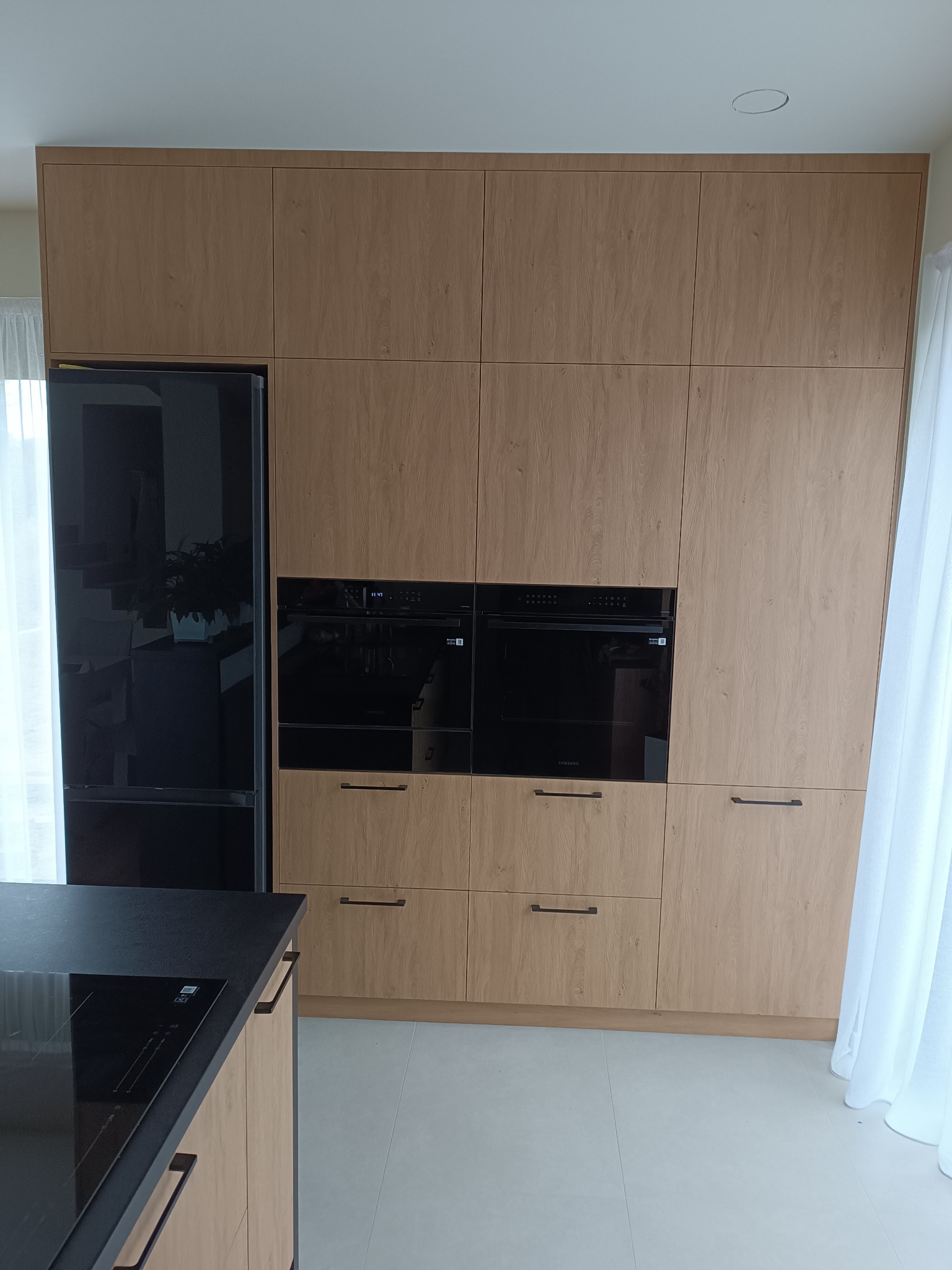 Jasna, drewnopodobna zabudowa kuchenna z czarnym sprzętem AGD (lodówka, piekarniki) i blatem. Minimalistyczne uchwyty. Ujęcie z dołu, widoczny fragment okna.