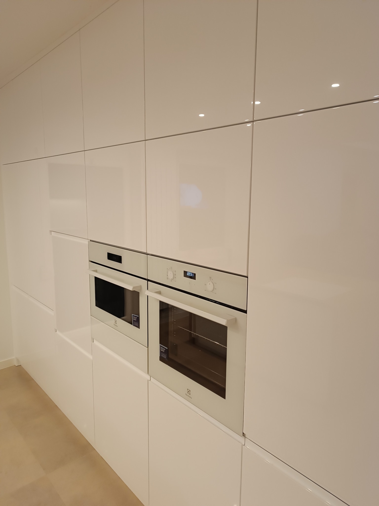 Biała zabudowa kuchenna z połyskiem, piekarnik i mikrofalówka Electrolux w zabudowie. Minimalistyczny design, gładkie fronty bez uchwytów.