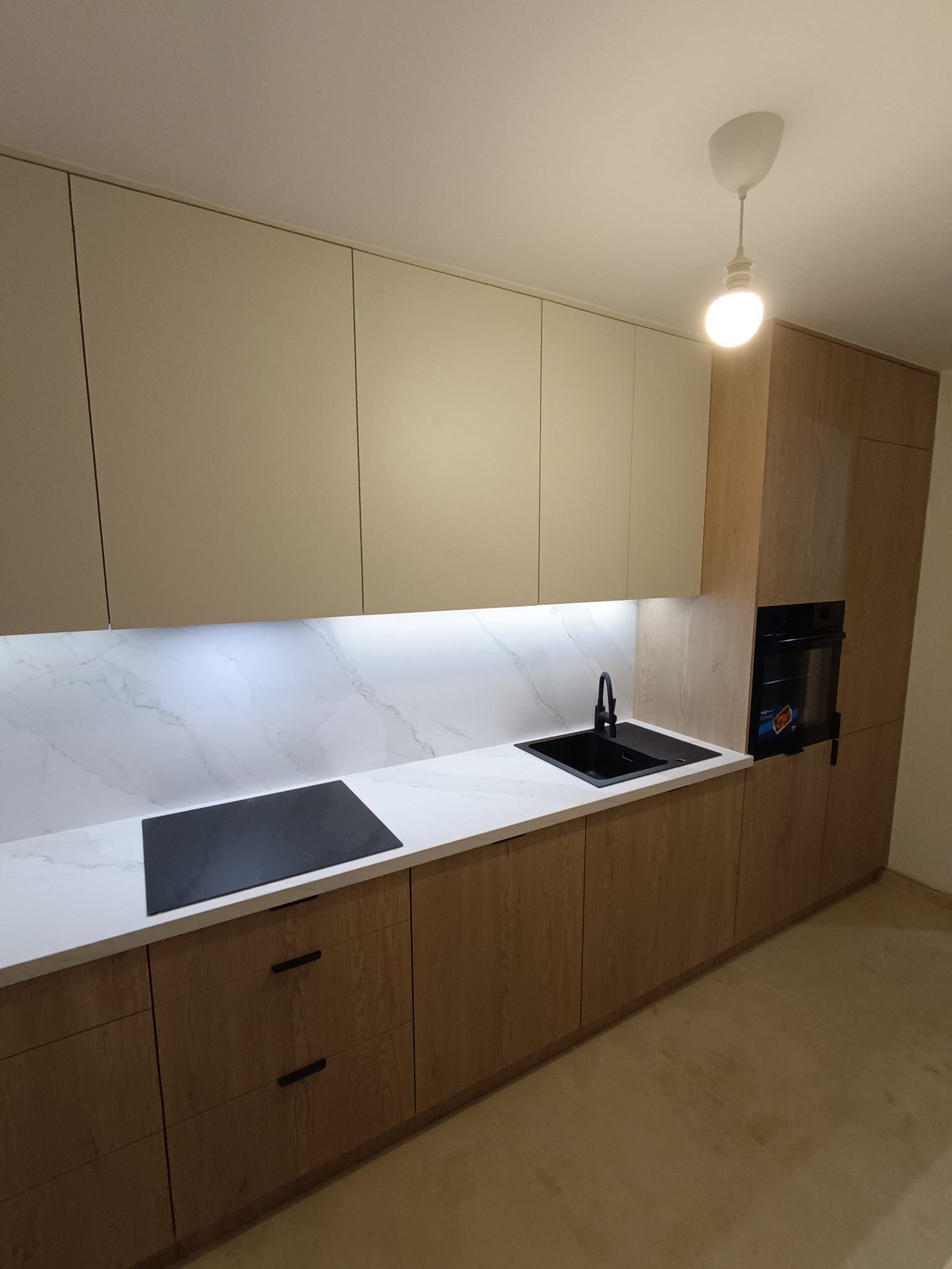 Nowoczesna kuchnia na wymiar: jasne górne szafki, drewnopodobne dolne, marmurowy blat z czarnym zlewem i płytą. Oświetlenie podszafkowe.