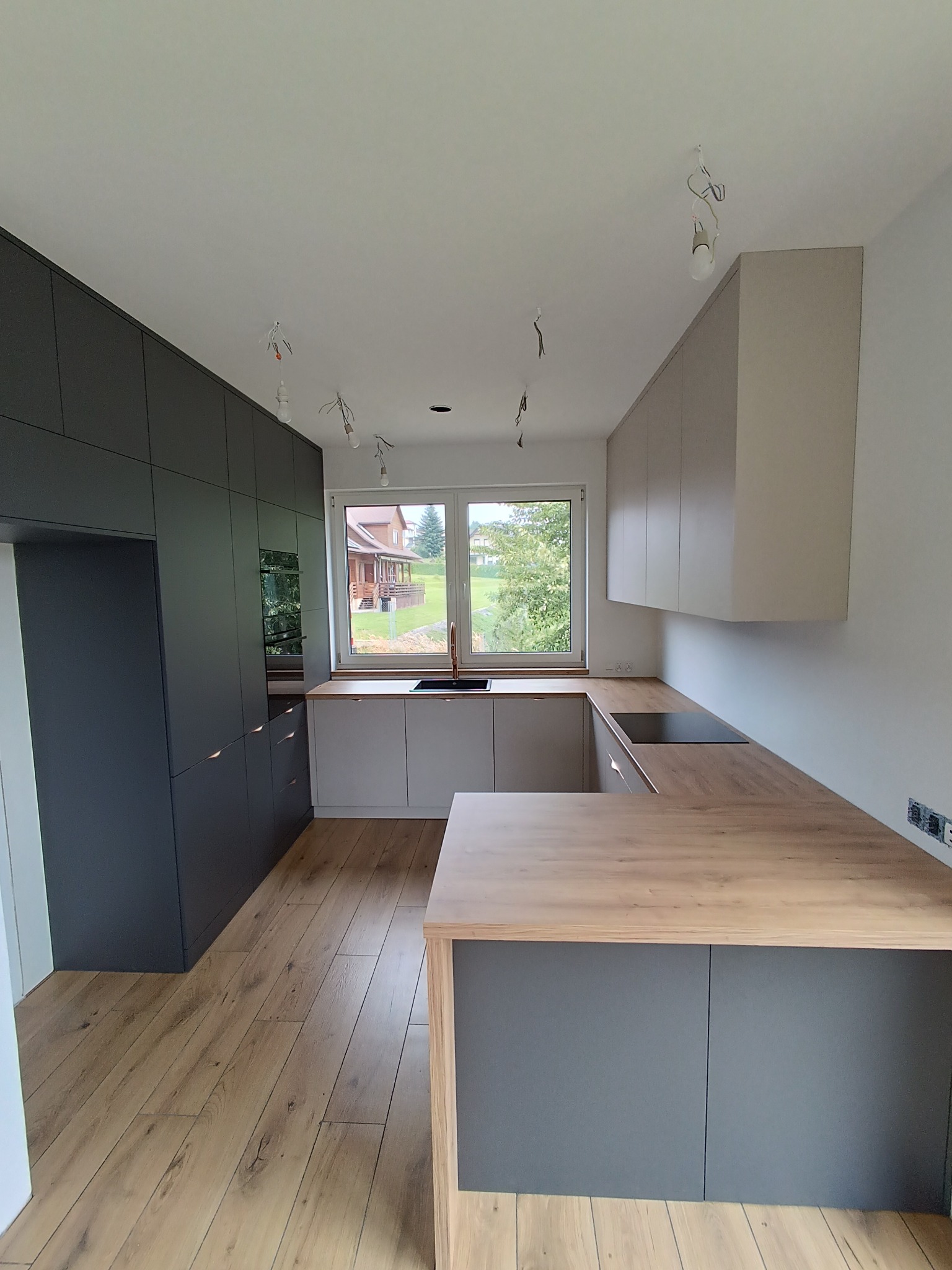 Nowoczesna kuchnia z drewnianym blatem i szarymi szafkami, widok na ogród przez okno. W tle widać niedokończone podłączenie oświetlenia.