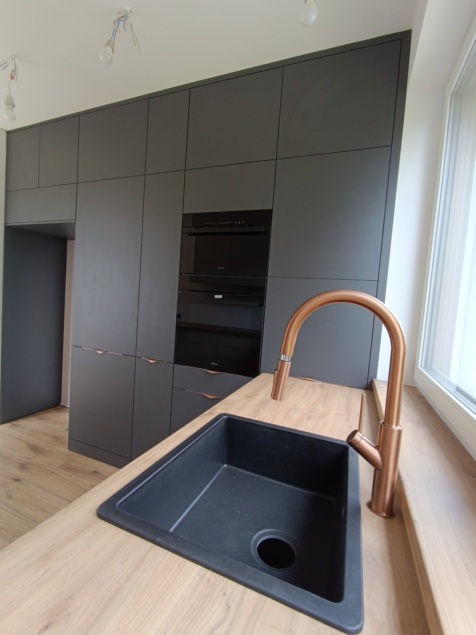 Nowoczesna kuchnia z szarymi frontami, drewnianym blatem i miedzianą baterią. W tle zabudowa z piekarnikiem i mikrofalówką. Niedokończone oświetlenie sufitowe.