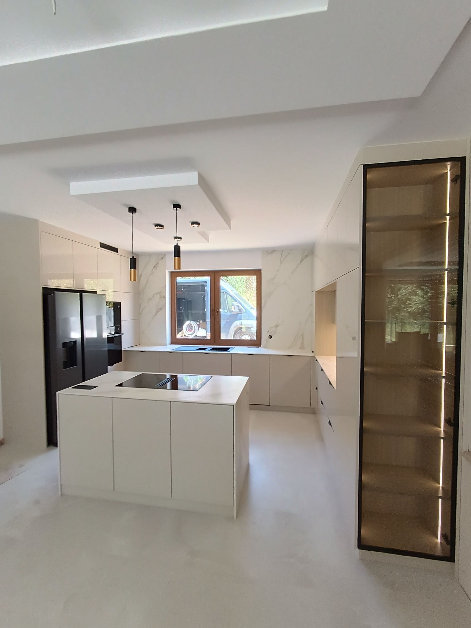 Jasna kuchnia z wyspą i czarną lodówką. Na ścianie marmurowy wzór, nad wyspą wiszą lampy. W tle okno, za nim zaparkowany van. Nowoczesny design.