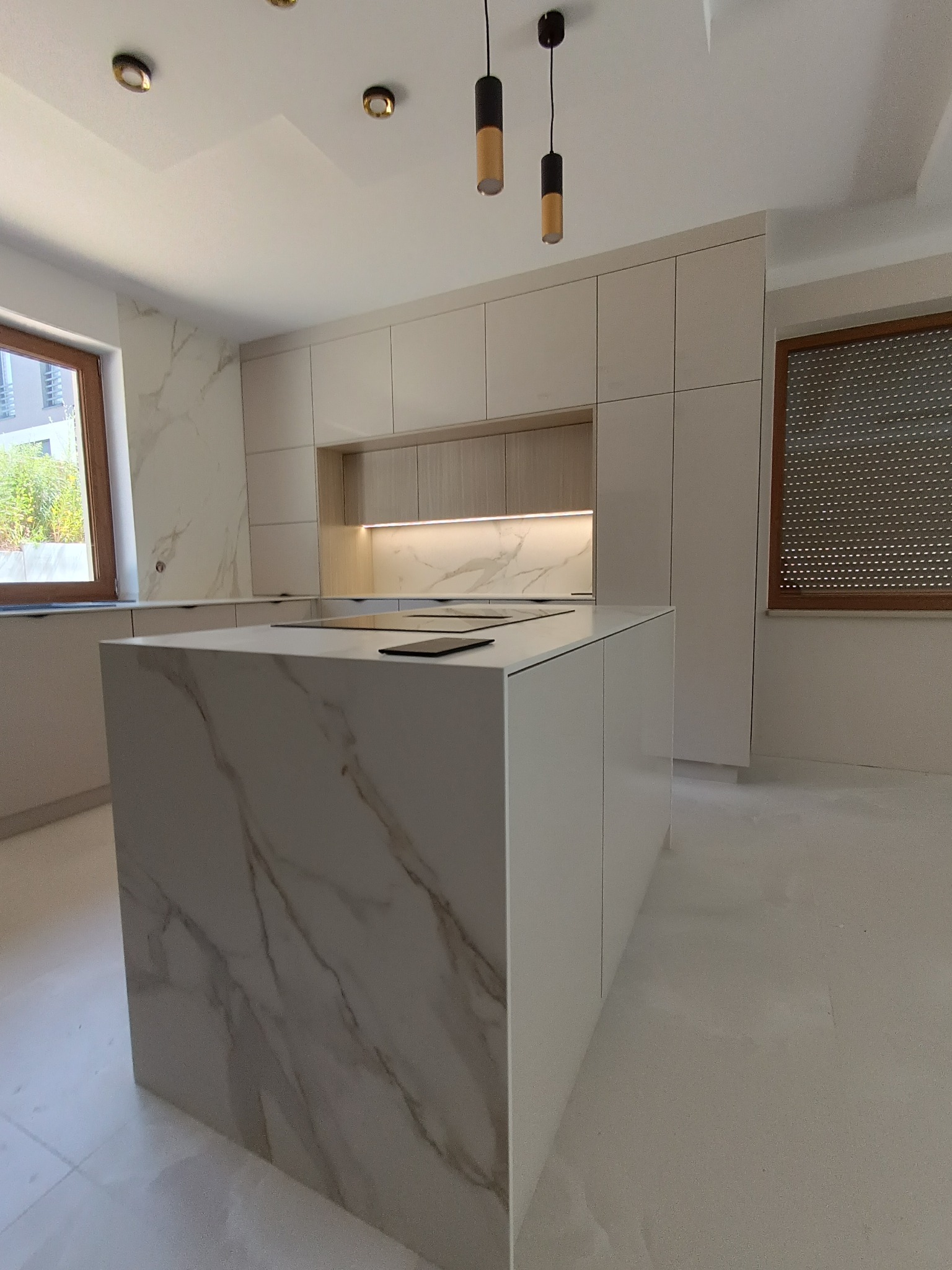 Nowoczesna kuchnia z wyspą, fronty w odcieniach bieli i beżu, marmurowy wzór na wyspie, minimalistyczne oświetlenie.