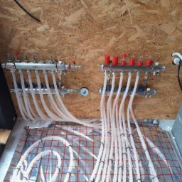 Rozdzielacz ogrzewania podłogowego z białymi rurkami na folii izolacyjnej z czerwoną siatką, zamontowany na ścianie z płyty OSB, widoczne zawory z czerwonymi pokrętłami i manometr.