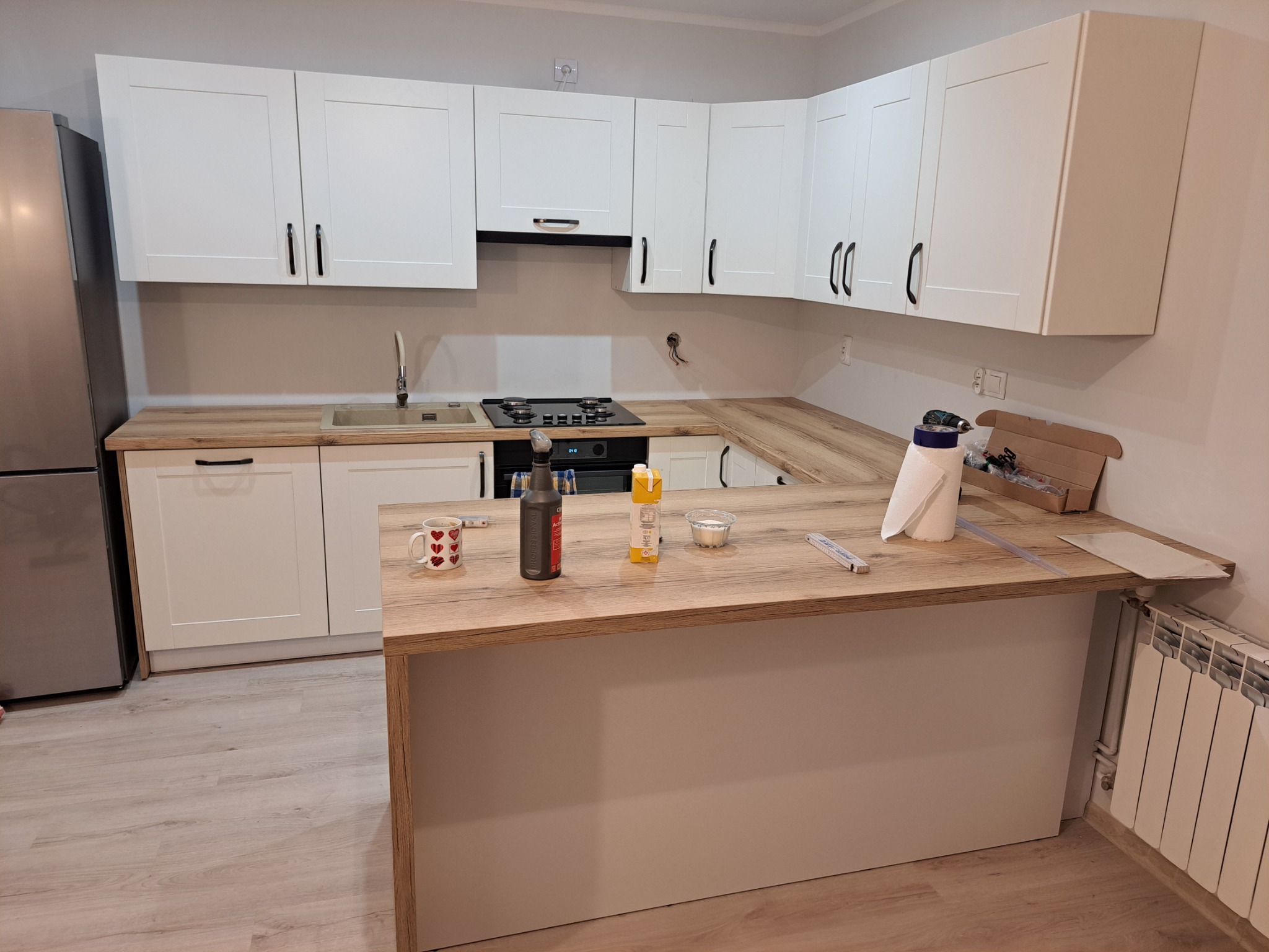 Nowoczesna kuchnia z białymi szafkami i blatem imitującym drewno, widoczne niedokończone prace wykończeniowe, takie jak wystające kable i brakujące listwy przypodłogowe. Na blacie stoi kubek...