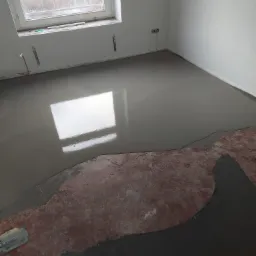 Wylewka samopoziomująca w trakcie rozprowadzania na podłożu, widoczna szpachelka i fragment starej podłogi, okno z widokiem na budynek