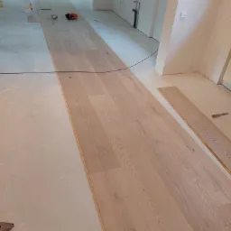 Układanie jasnego parkietu w przestronnym wnętrzu, widoczne narzędzia i niedokończona podłoga.