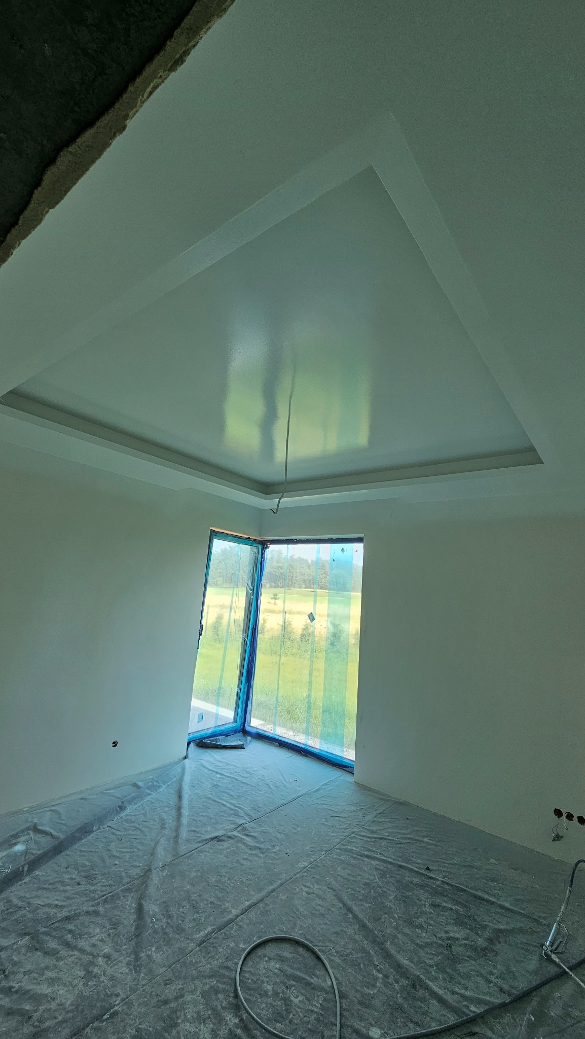 Przygotowane do malowania wnętrze z zabezpieczoną folią podłogą, świeżo wykończone gładzie na ścianach i suficie z dekoracyjnym obniżeniem, okno zabezpieczone folią malarską.