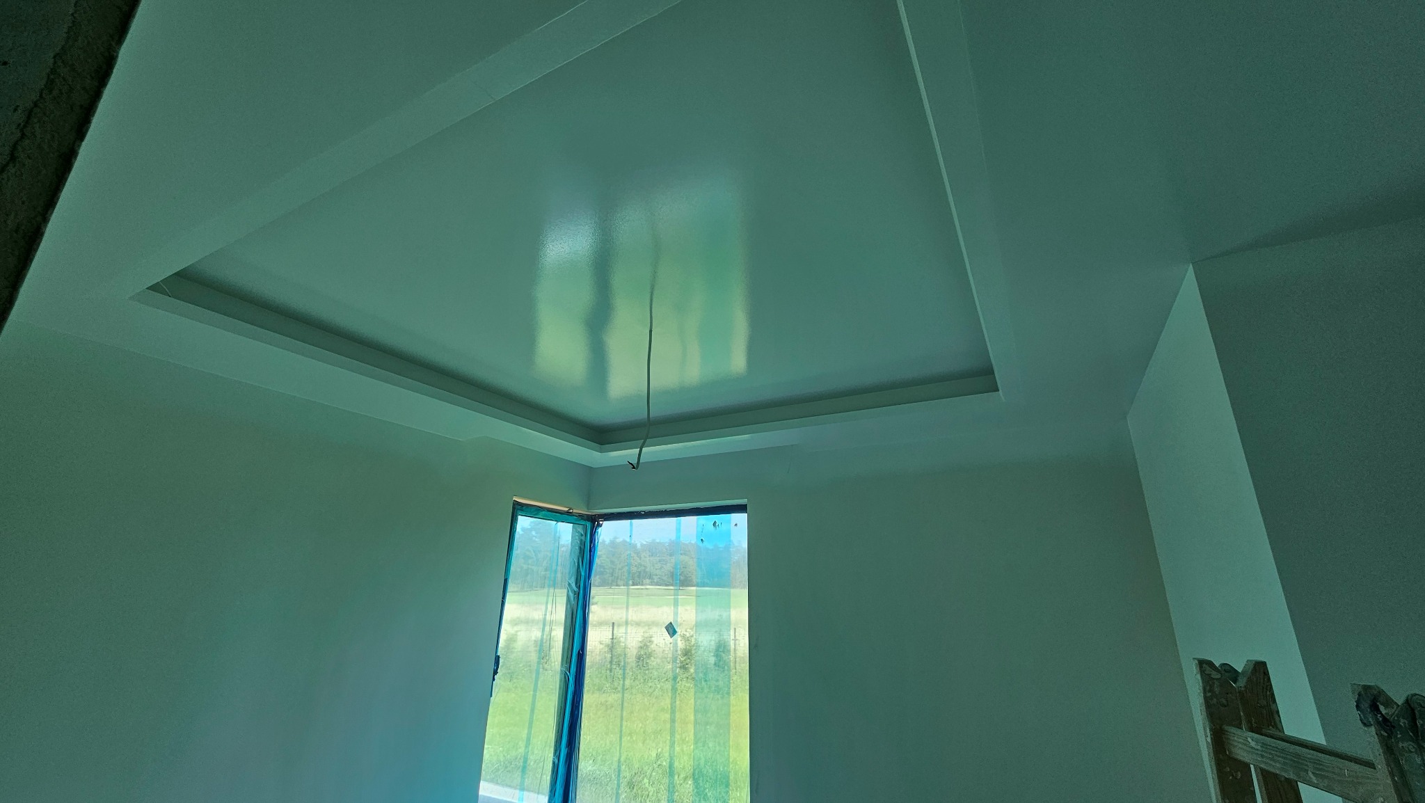 Świeżo pomalowane wnętrze z podwieszanym sufitem, widoczne odbicia światła, okno zabezpieczone folią malarską, drabina w rogu.