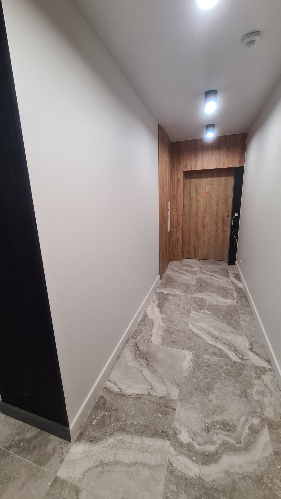 Korytarz po remoncie z białymi ścianami, szarą marmurową podłogą i drewnianą okładziną ściany z drzwiami wejściowymi
