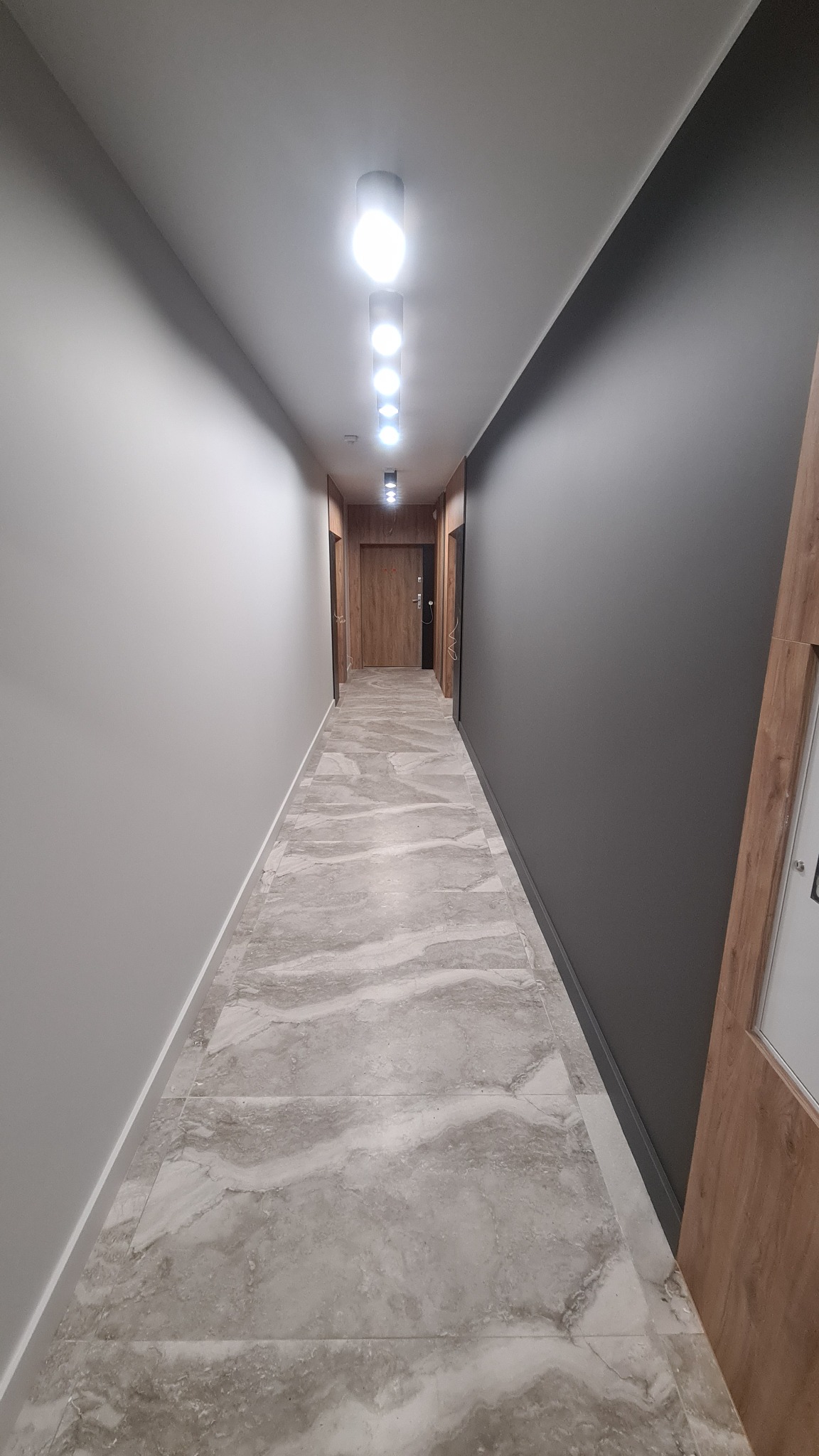 Korytarz w Gdyni po remoncie: ściany pomalowane na szaro i biało, marmurowe płytki na podłodze, oświetlenie sufitowe w postaci rzędu lamp.