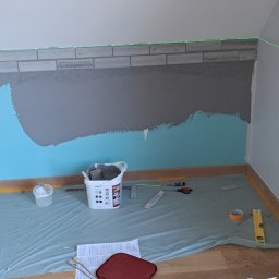 Damian Konopka - Ściana w trakcie wykańczania z położonymi płytkami imitującymi cegłę, zabezpieczona folią podłoga i narzędzia w pomieszczeniu ze skośnym sufitem.