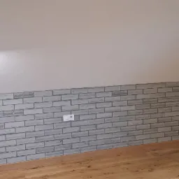 Poddasze po remoncie: ściana wykończona szarą cegłą dekoracyjną, drewniana podłoga, widoczny uchwyt do telewizora i gniazdka elektryczne.