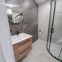 Damian Konopka - Nowoczesna łazienka z umywalką na szafce z drewnianym frontem, okrągłym lustrem i kabiną prysznicową z czarnymi profilami na tle marmurowych płytek.