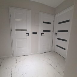 Damian Konopka - Trzy białe, nowoczesne drzwi wewnętrzne z czarnymi akcentami na tle jasnych ścian i podłogi imitującej marmur. Minimalistyczny design, widoczne klamki i listwy przypodłogowe.