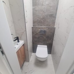 Damian Konopka - Wykończona łazienka w odcieniach bieli i szarości z marmurkowymi płytkami, wiszącą toaletą i umywalką z drewnianą szafką oraz czarną armaturą, widziana z perspektywy wejścia.