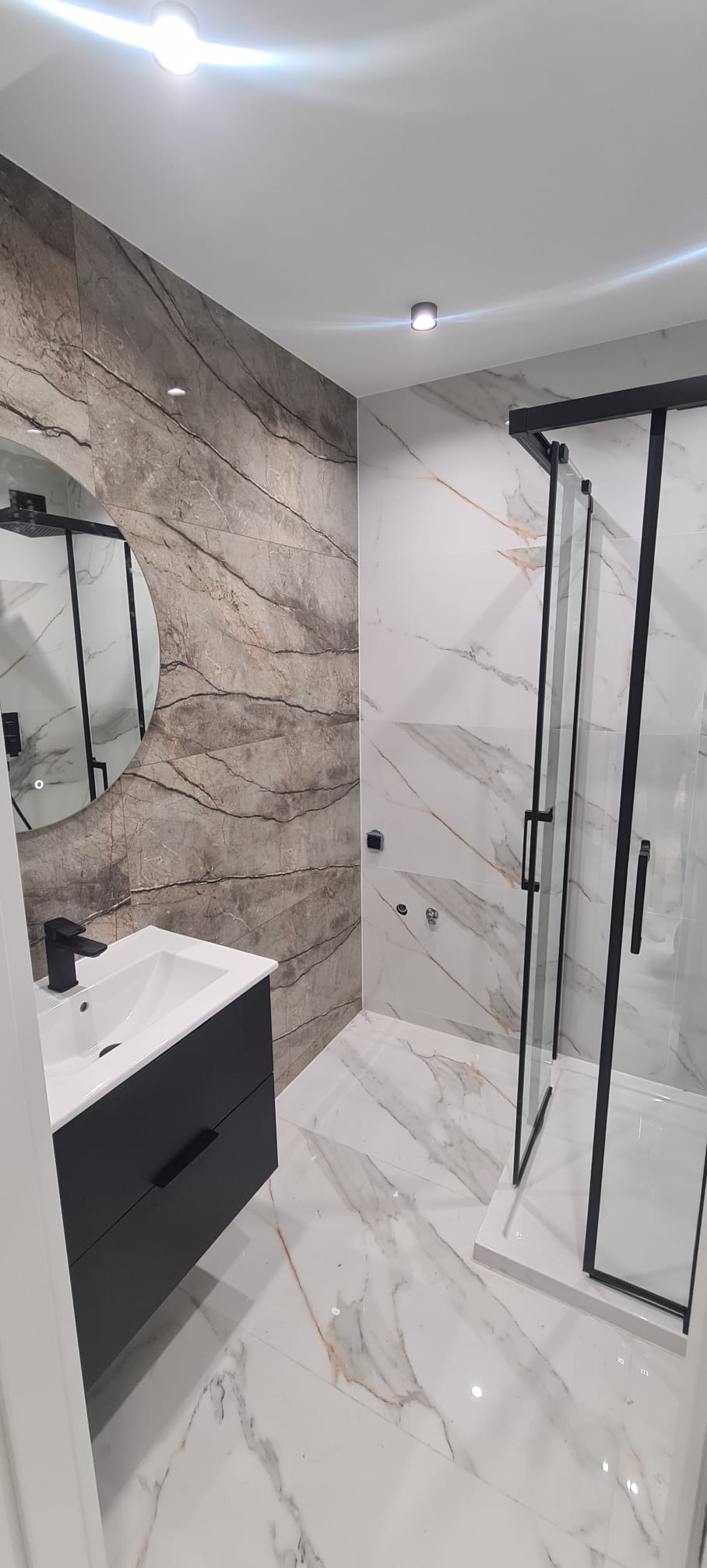 Nowoczesna łazienka z marmurowymi płytkami, czarną armaturą i kabiną prysznicową typu walk-in, oświetlona punktowymi lampami sufitowymi.