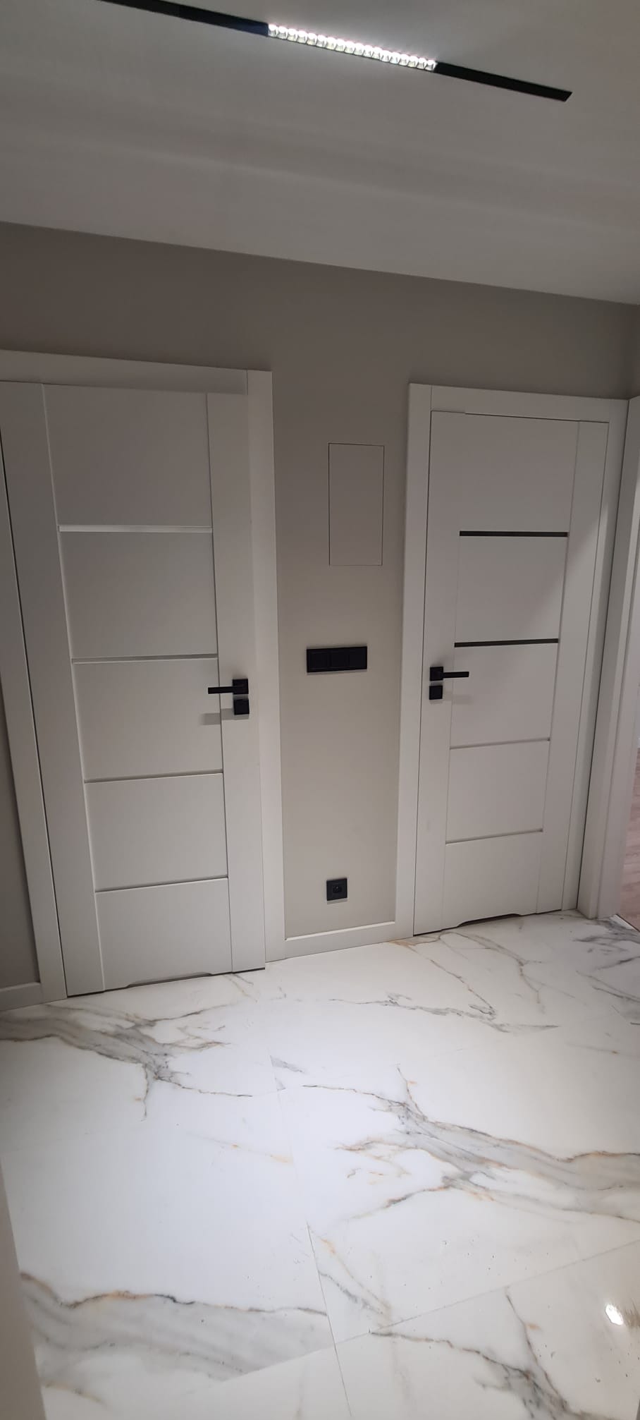 Dwa białe, nowoczesne drzwi wewnętrzne z czarnymi klamkami na tle marmurowej podłogi i szarej ściany z czarnymi włącznikami.