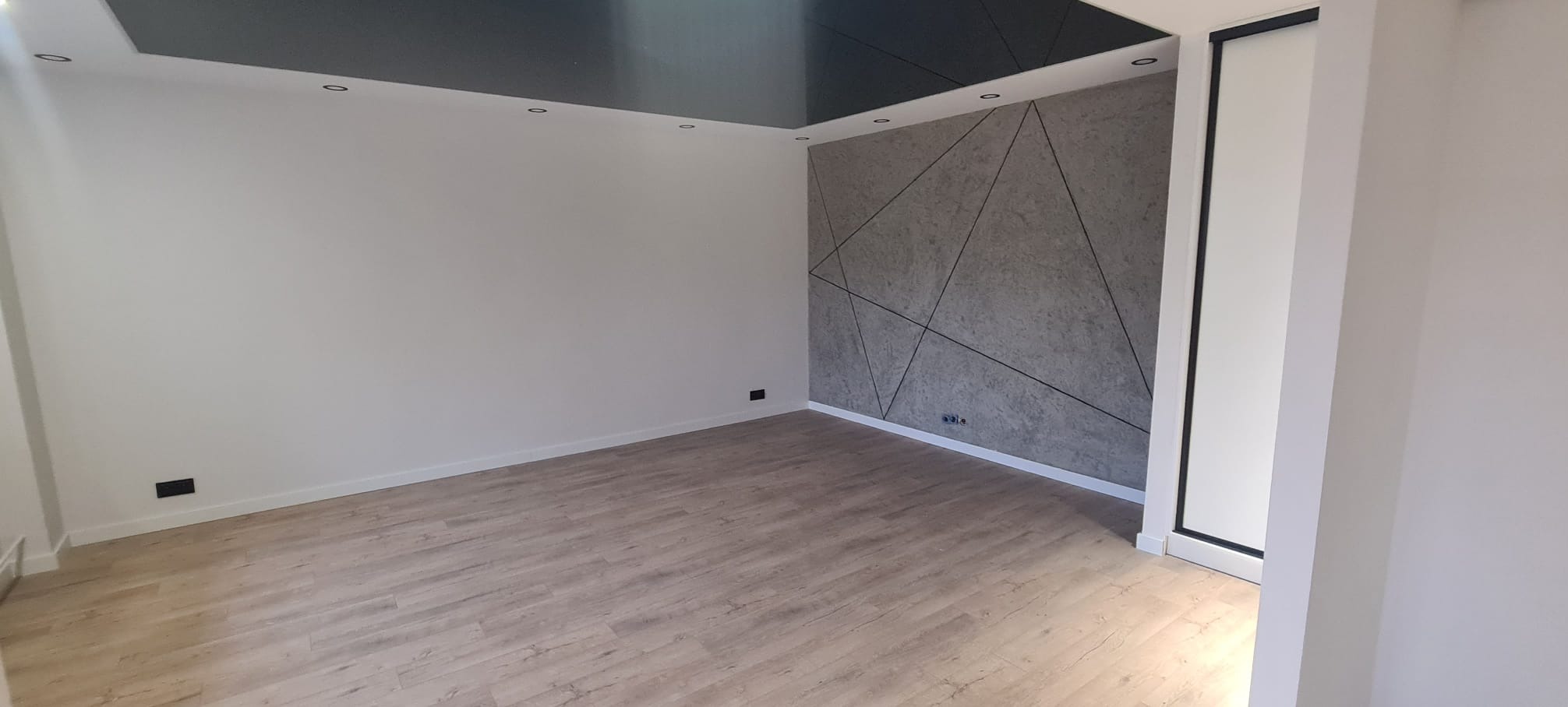 Wnętrze pokoju po remoncie z panelami ściennymi w geometryczne wzory, parkietem i podwieszanym sufitem z oświetleniem punktowym.