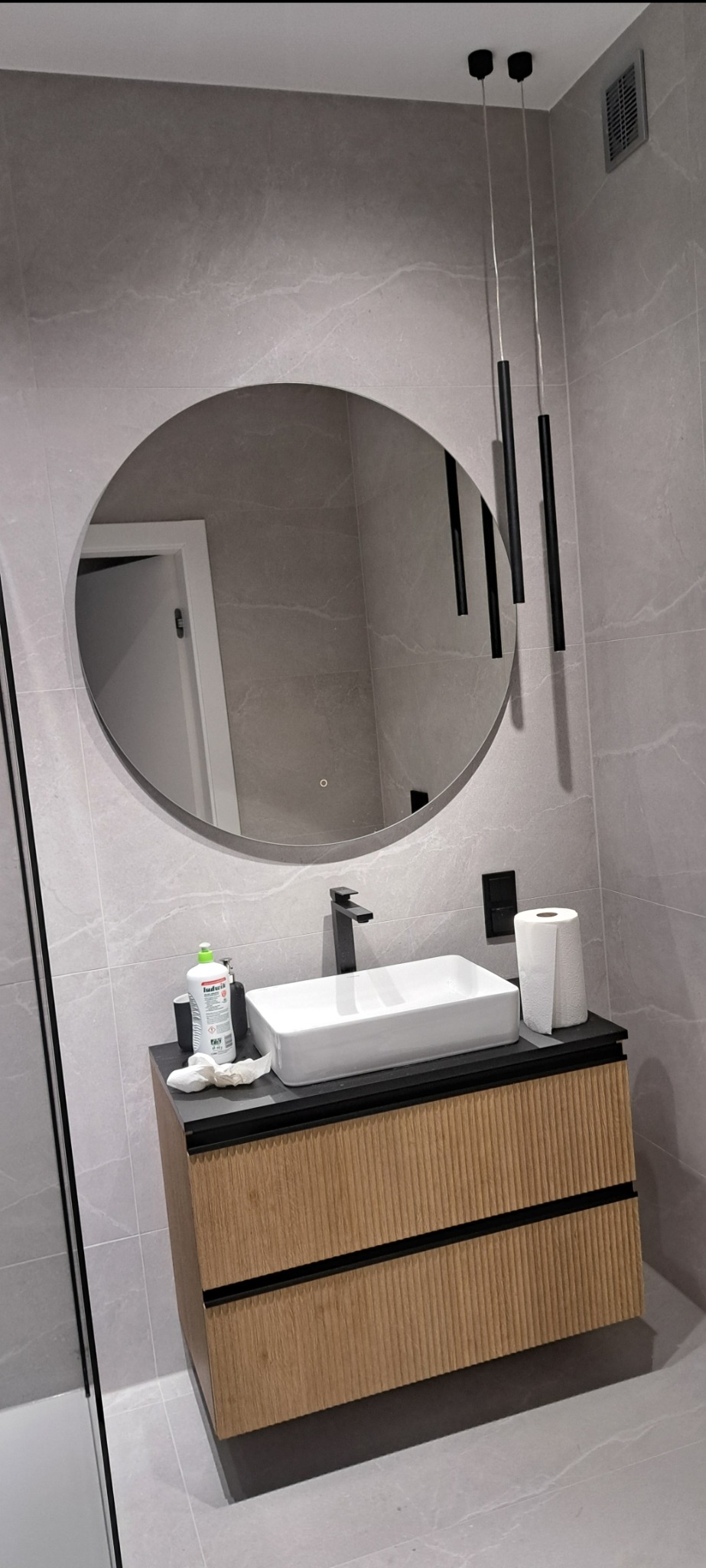 Nowoczesna łazienka z okrągłym lustrem, wiszącymi lampami, prostokątną umywalką nablatową na szafce z drewnianymi lamelami i szarą marmurową okładziną ścienną.