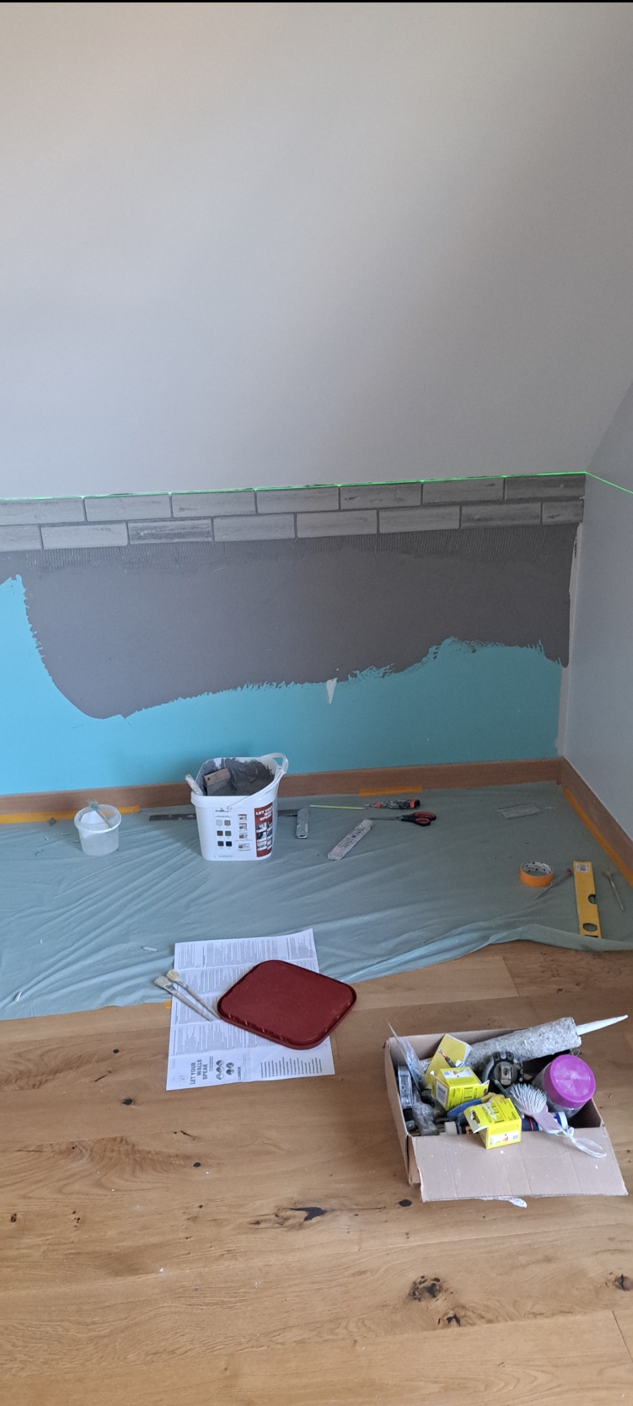 Ściana w trakcie wykańczania z położonymi płytkami imitującymi cegłę, zabezpieczona folią podłoga i narzędzia w pomieszczeniu ze skośnym sufitem.