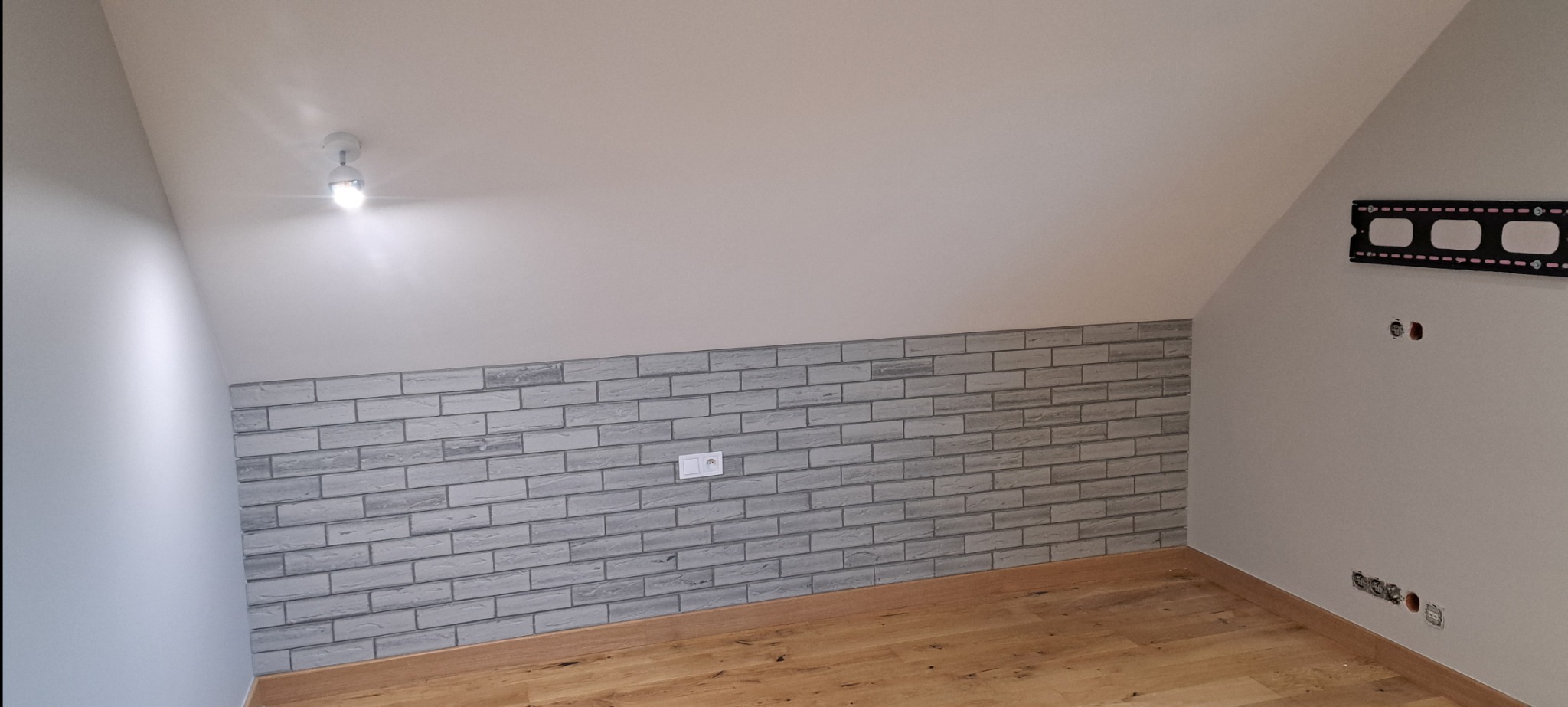 Poddasze po remoncie: ściana wykończona szarą cegłą dekoracyjną, drewniana podłoga, widoczny uchwyt do telewizora i gniazdka elektryczne.