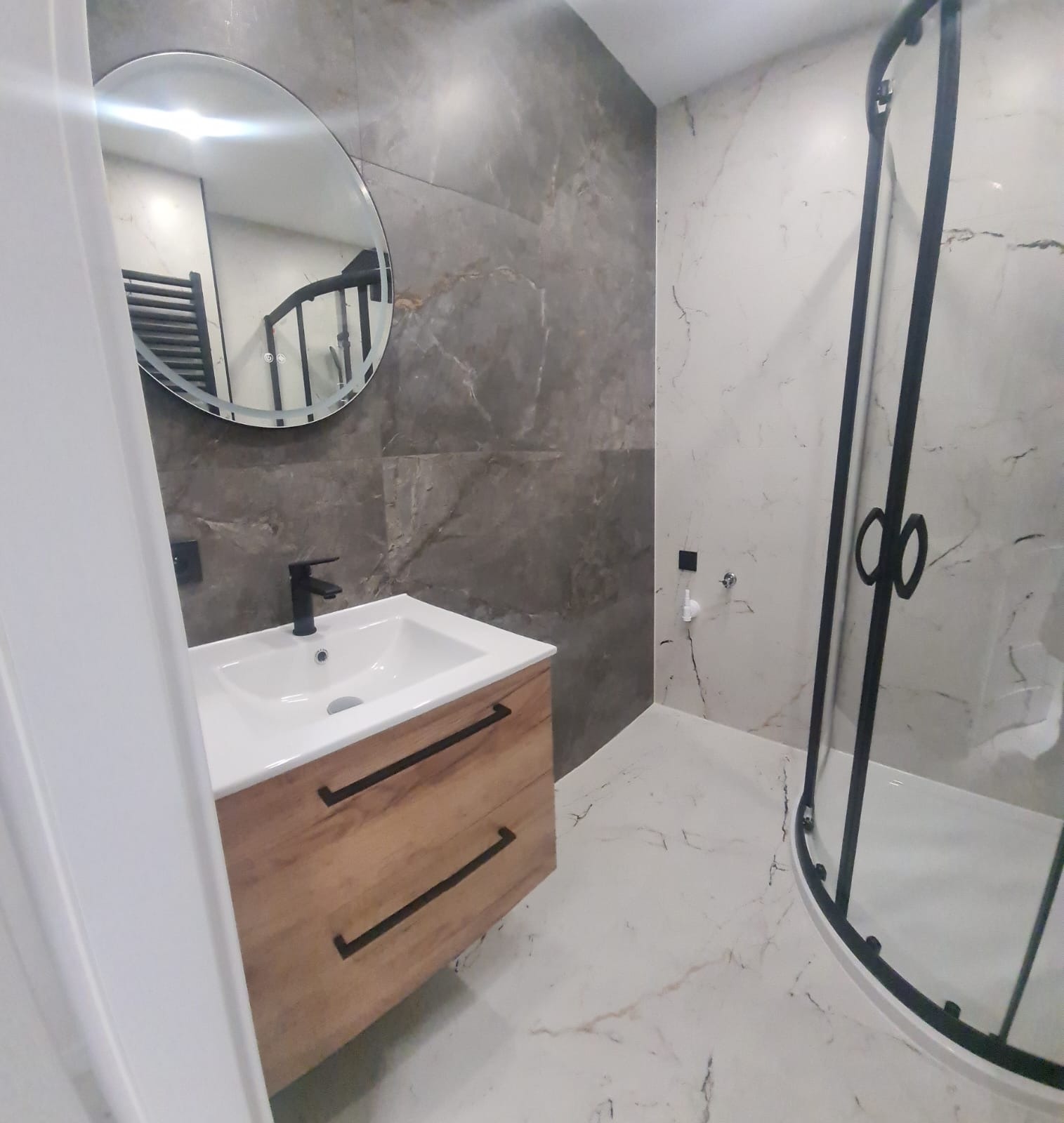 Nowoczesna łazienka z umywalką na szafce z drewnianym frontem, okrągłym lustrem i kabiną prysznicową z czarnymi profilami na tle marmurowych płytek.