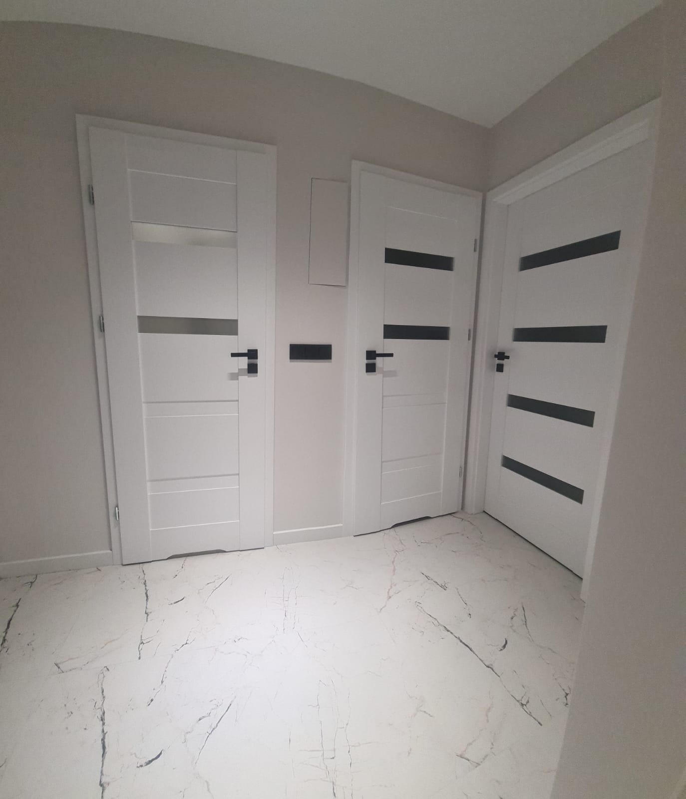 Trzy białe, nowoczesne drzwi wewnętrzne z czarnymi akcentami na tle jasnych ścian i podłogi imitującej marmur. Minimalistyczny design, widoczne klamki i listwy przypodłogowe.