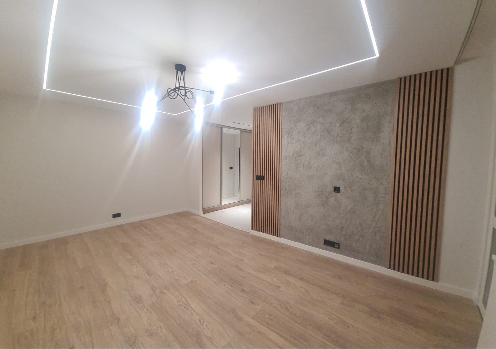 Nowoczesne wnętrze po remoncie: drewniana podłoga, ściana z dekoracyjnym betonem i drewnianymi lamelami, oświetlenie LED na suficie. Minimalistyczny design.