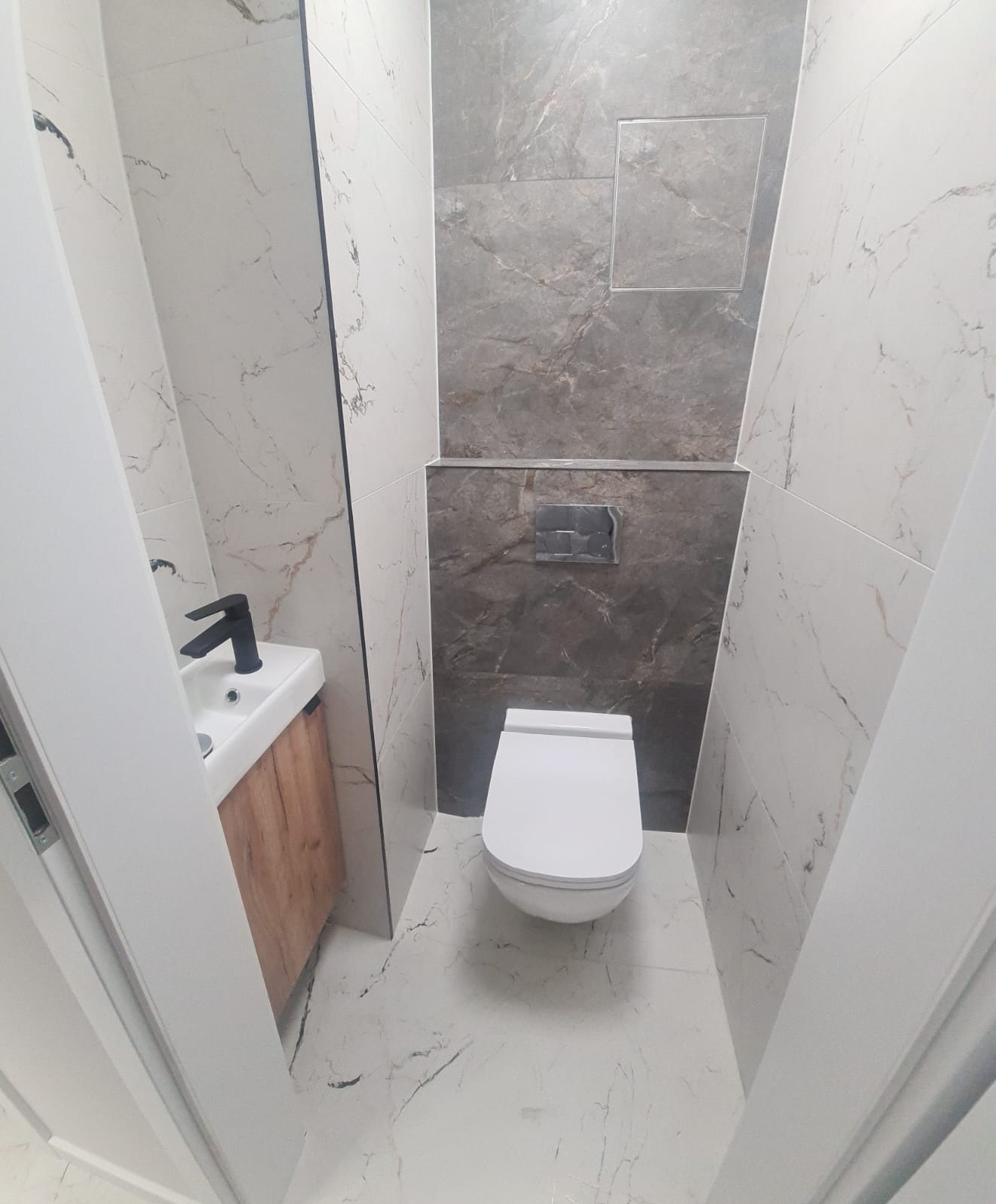 Wykończona łazienka w odcieniach bieli i szarości z marmurkowymi płytkami, wiszącą toaletą i umywalką z drewnianą szafką oraz czarną armaturą, widziana z perspektywy wejścia.