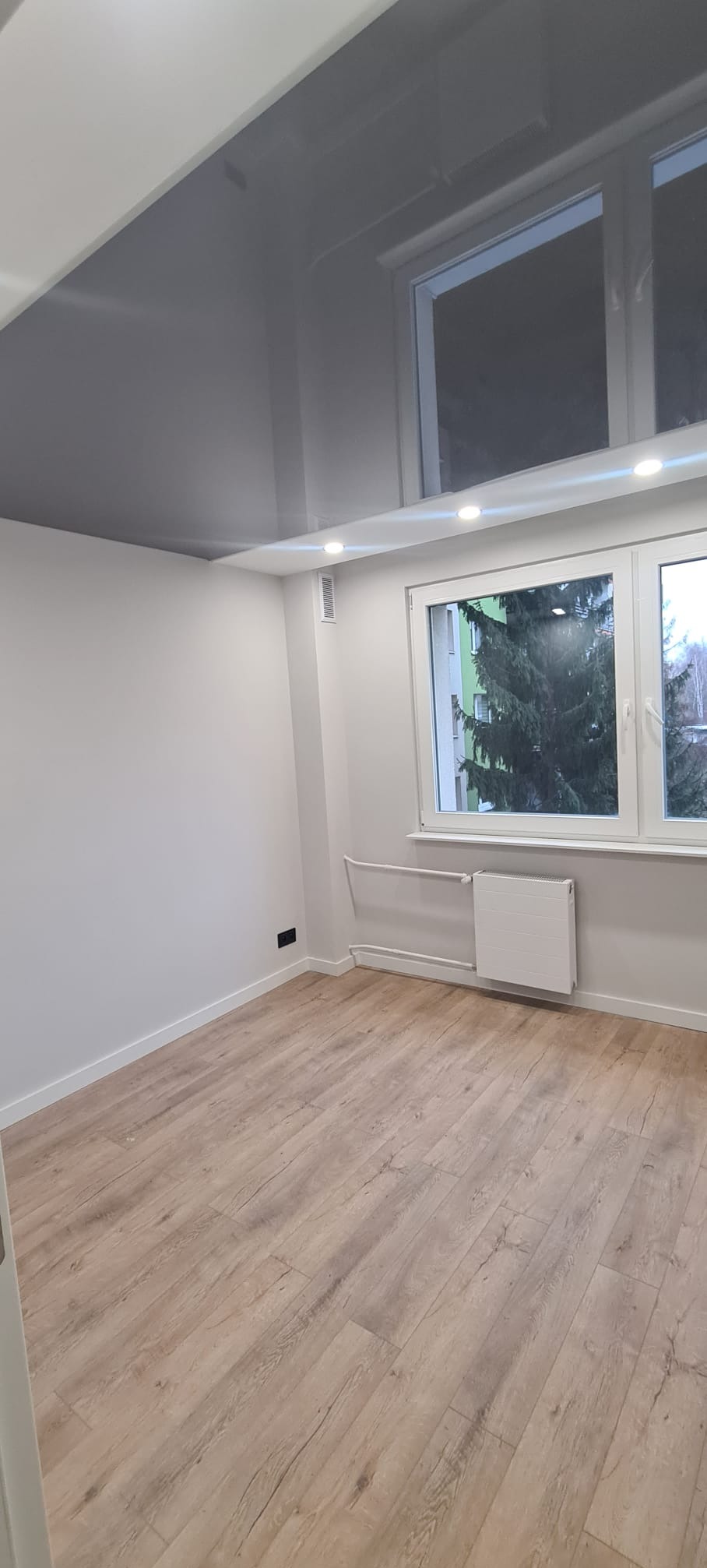 Jasne panele podłogowe w pokoju z białymi ścianami i błyszczącym, szarym sufitem napinanym. Widoczne okno z grzejnikiem i fragment drzewa za oknem.