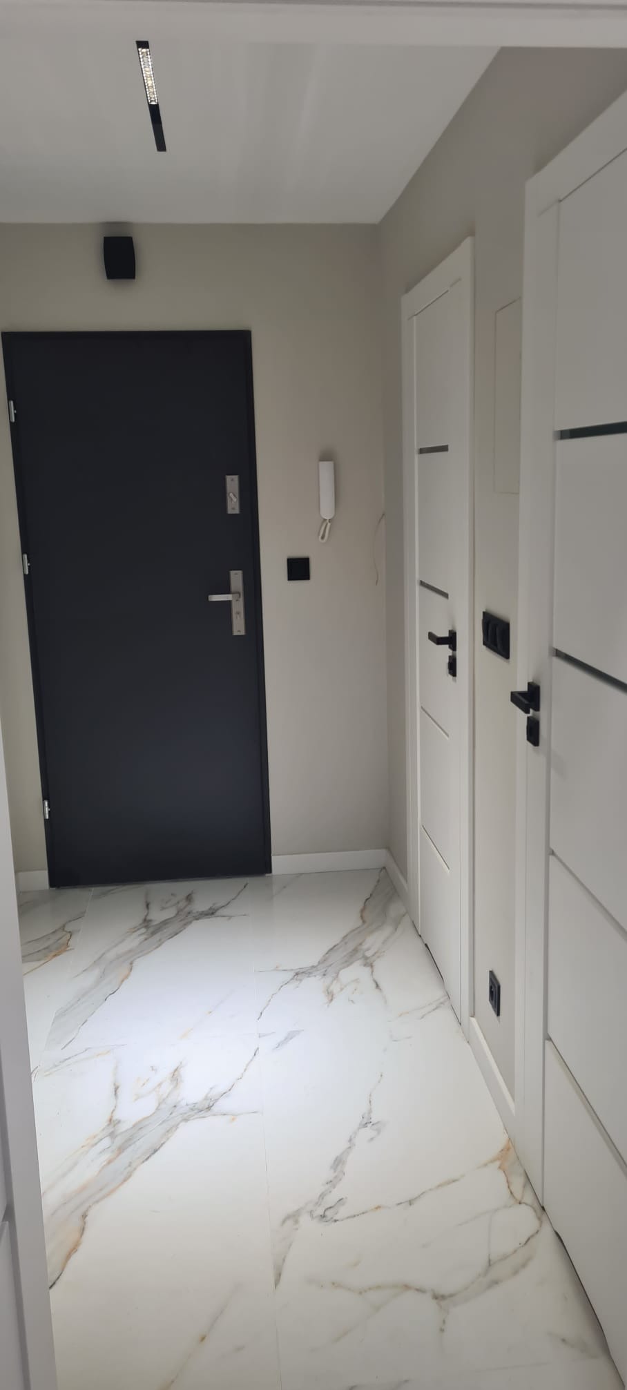 Nowoczesny przedpokój z czarnymi drzwiami wejściowymi i białymi drzwiami wewnętrznymi. Podłoga wyłożona białymi płytkami imitującymi marmur z delikatnym złotym żyłowaniem.