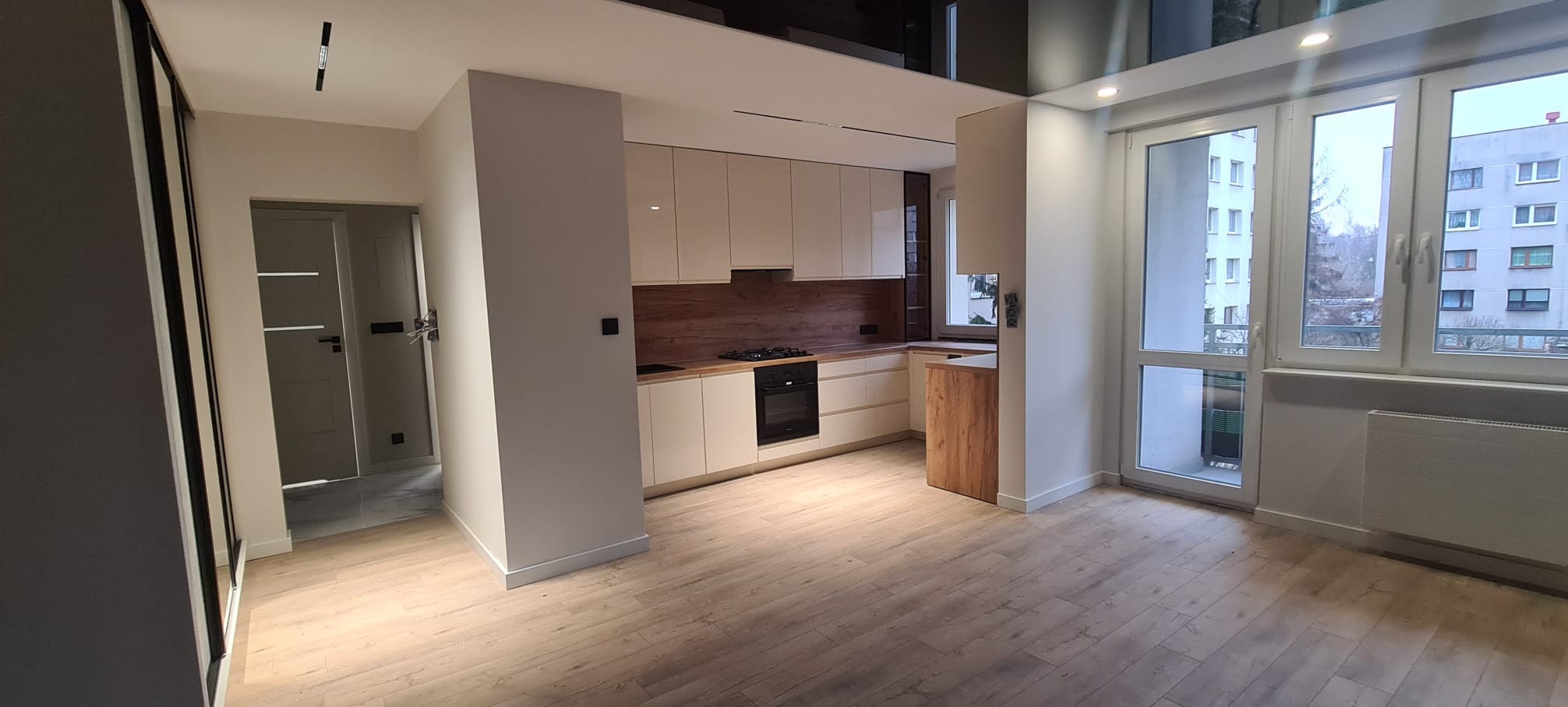 Jasna kuchnia z drewnianymi akcentami i parkietem, widok na balkon przez przeszklone drzwi. Minimalistyczny design, nowoczesne oświetlenie punktowe.