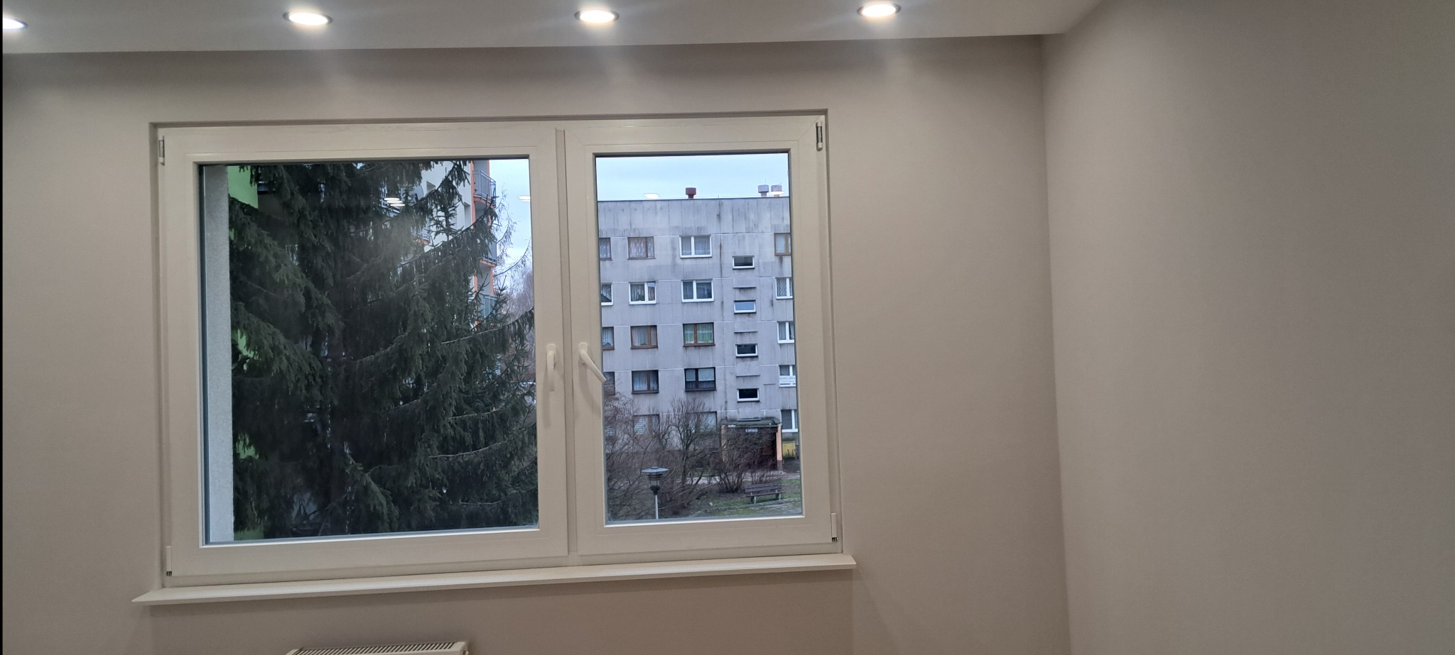 Jasne, świeżo pomalowane wnętrze z białym oknem, przez które widać budynek mieszkalny i drzewo. Minimalistyczny wystrój, neutralne barwy ścian i sufitu.