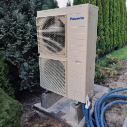 Zewnętrzna jednostka klimatyzacji Panasonic zamontowana na betonowych podstawach w ogrodzie, z widocznymi niebieskimi przewodami instalacyjnymi.